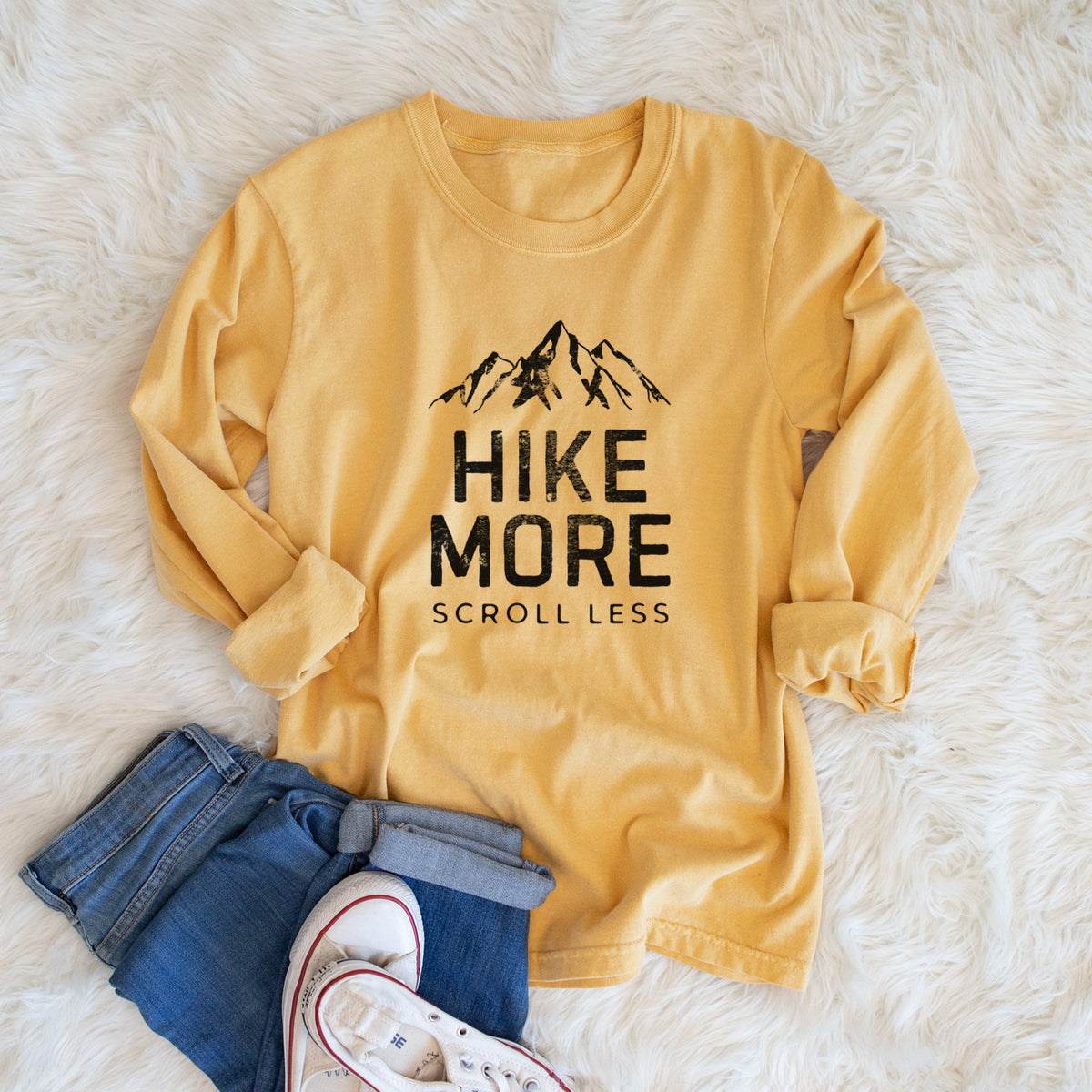 Golden wheat yellow Comfort Colors long sleeve shirt printed with black mountain peaks above the bold words Hike More, Scroll Less, shown in a lifestyle photo.