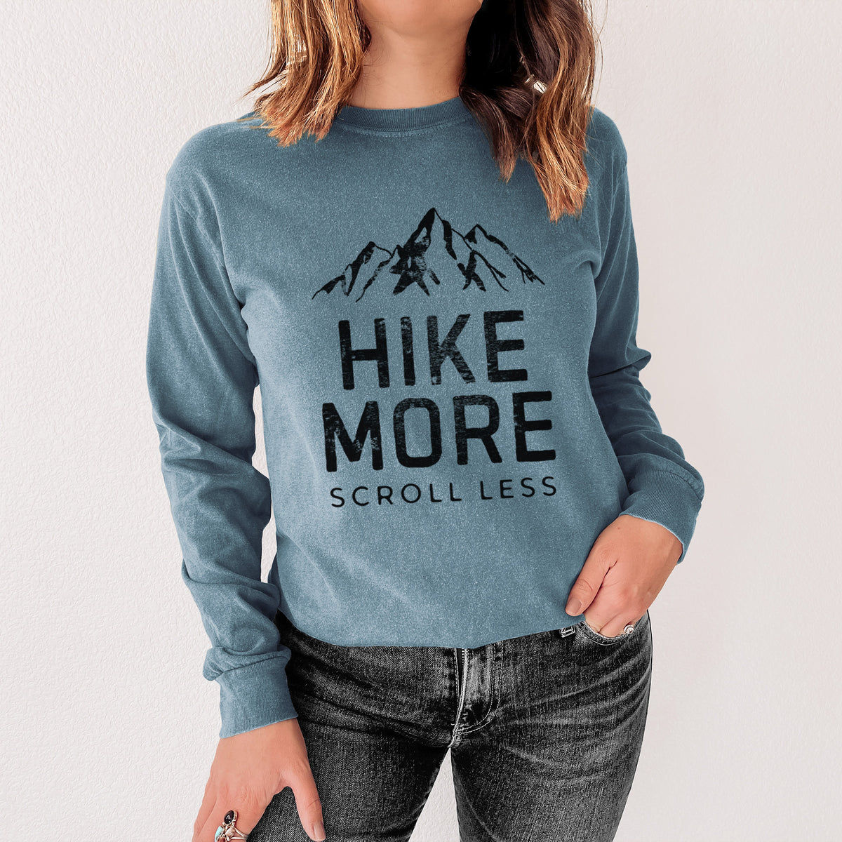 Steel blue Comfort Colors long sleeve shirt printed with black mountain peaks above the bold words Hike More, Scroll Less, shown in a lifestyle photo.