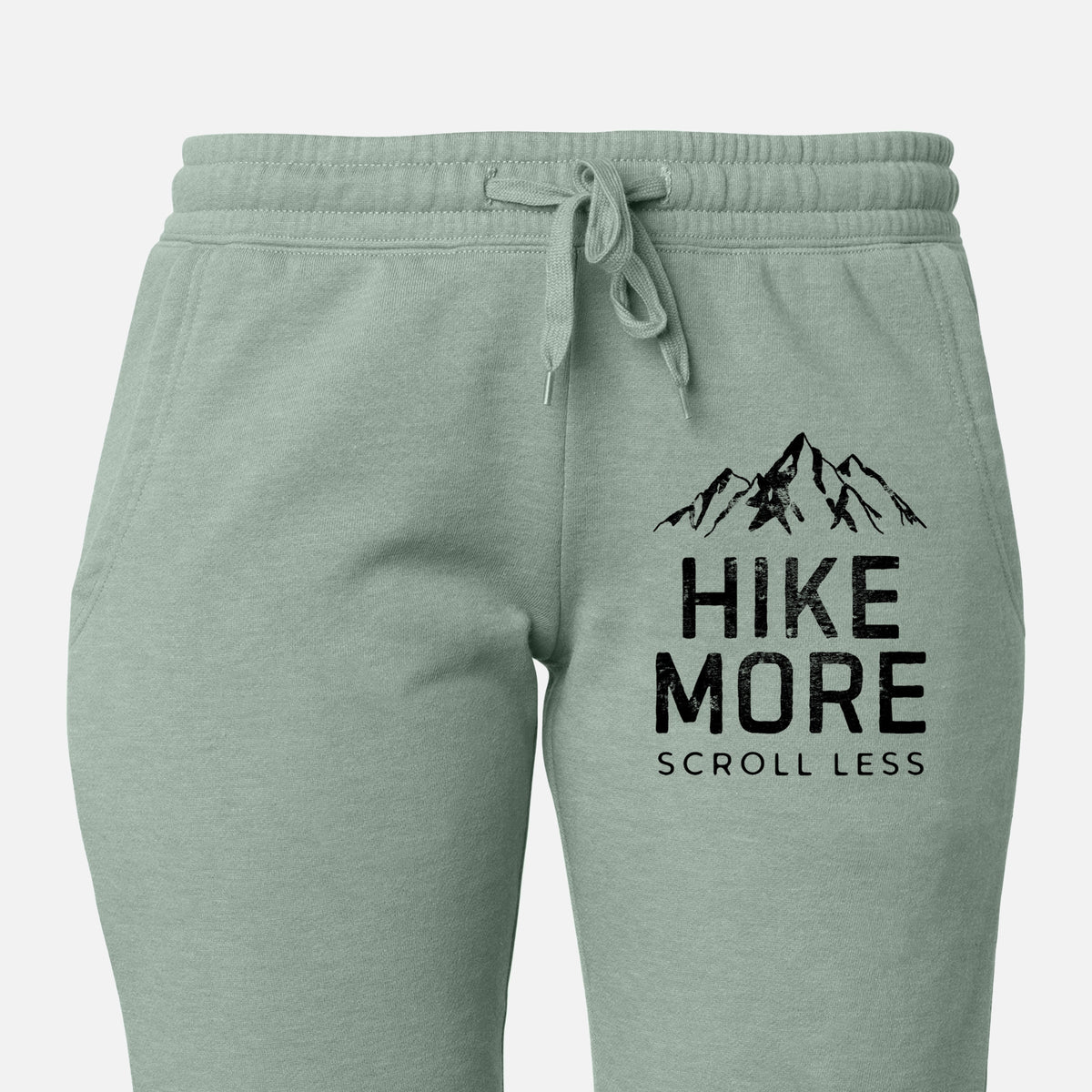 Front of sage green Cali Wave sweatpants, printed with black mountain peaks above the bold words Hike More, Scroll Less on the leg.