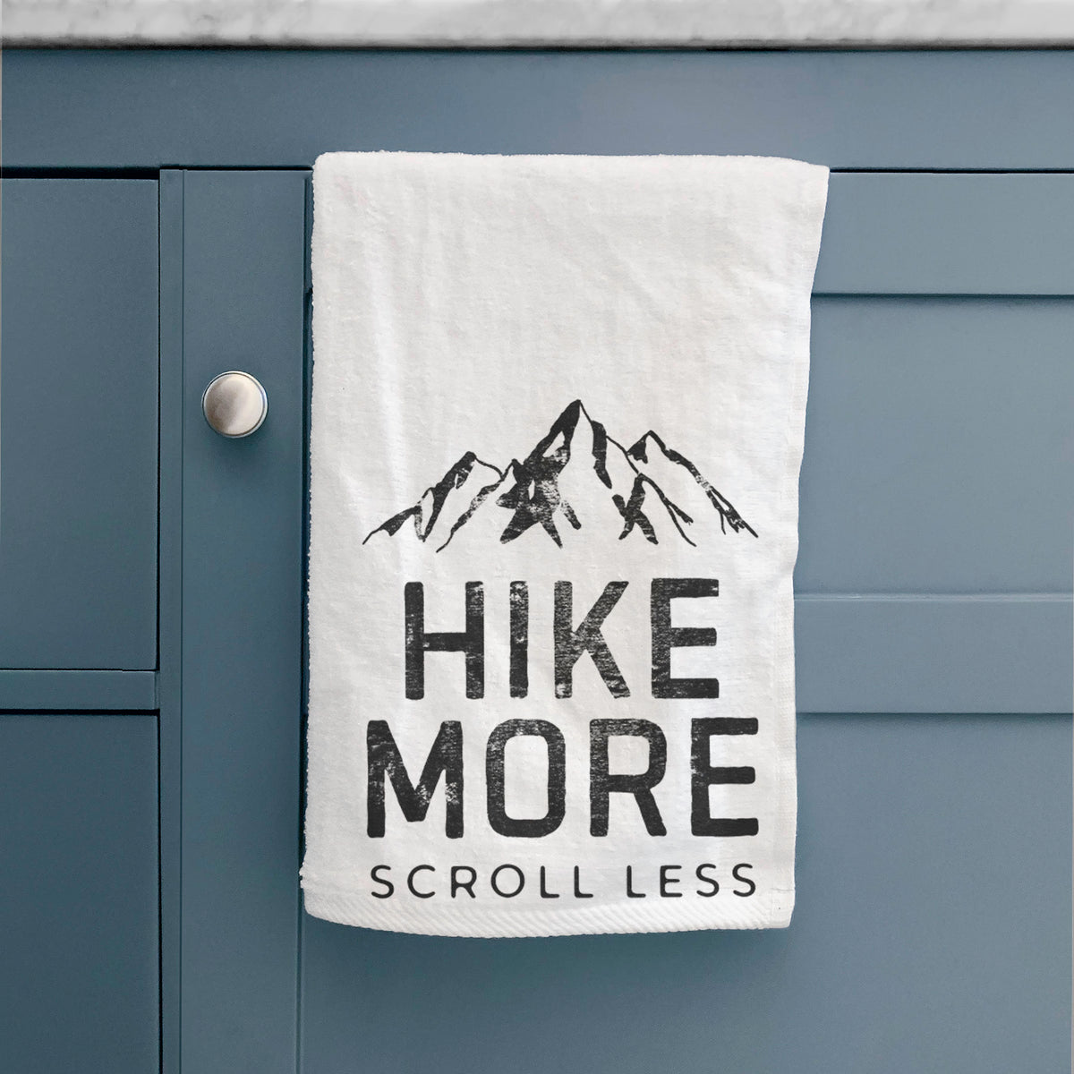 White hand towel printed with black mountain peaks above the bold words Hike More, Scroll Less, hanging on a cabinet door.