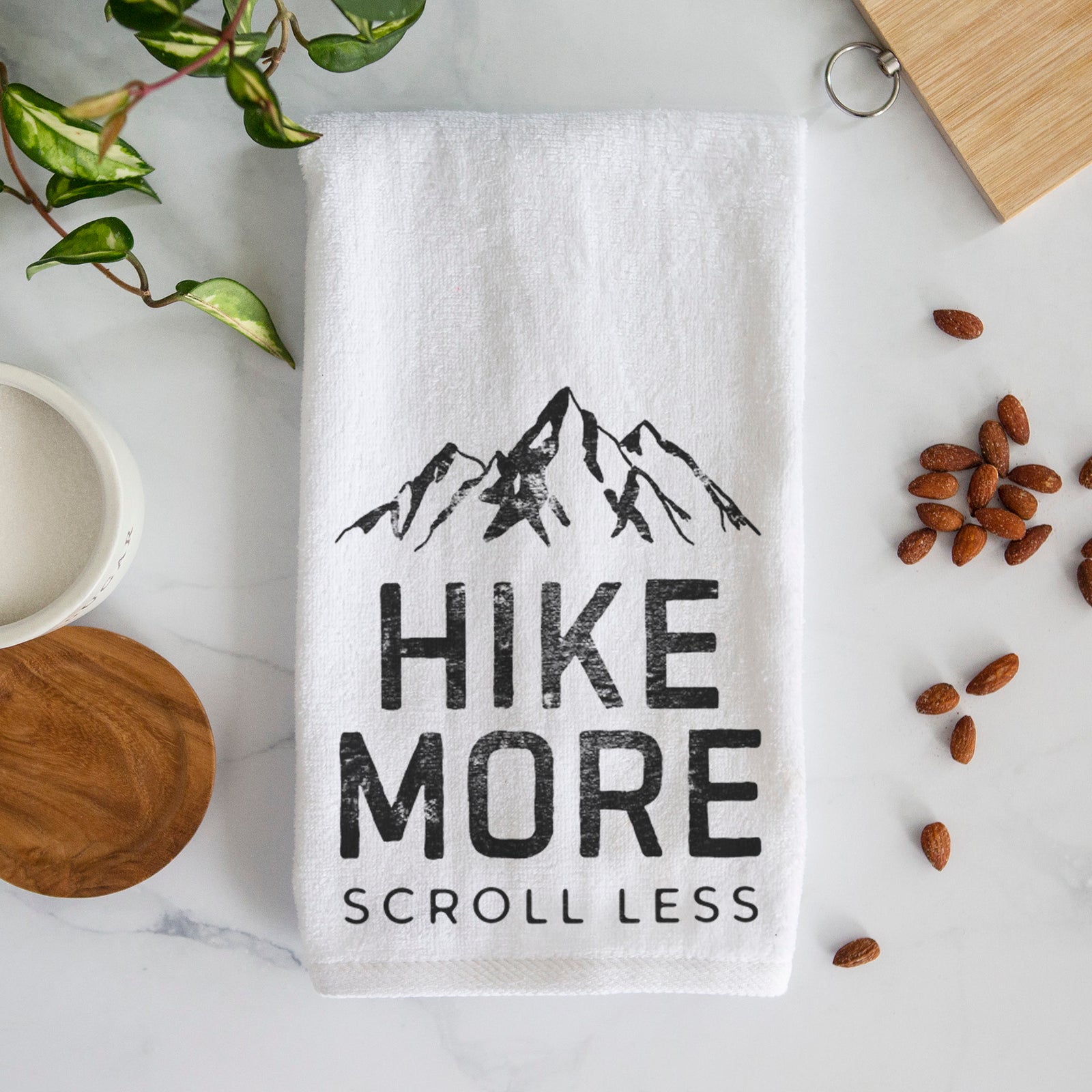 White hand towel printed with black mountain peaks above the bold words Hike More, Scroll Less.
