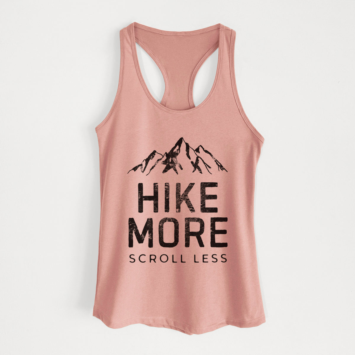 Desert pink women's racerback tank top laid flat, printed with black mountain peaks above the bold words Hike More, Scroll Less.