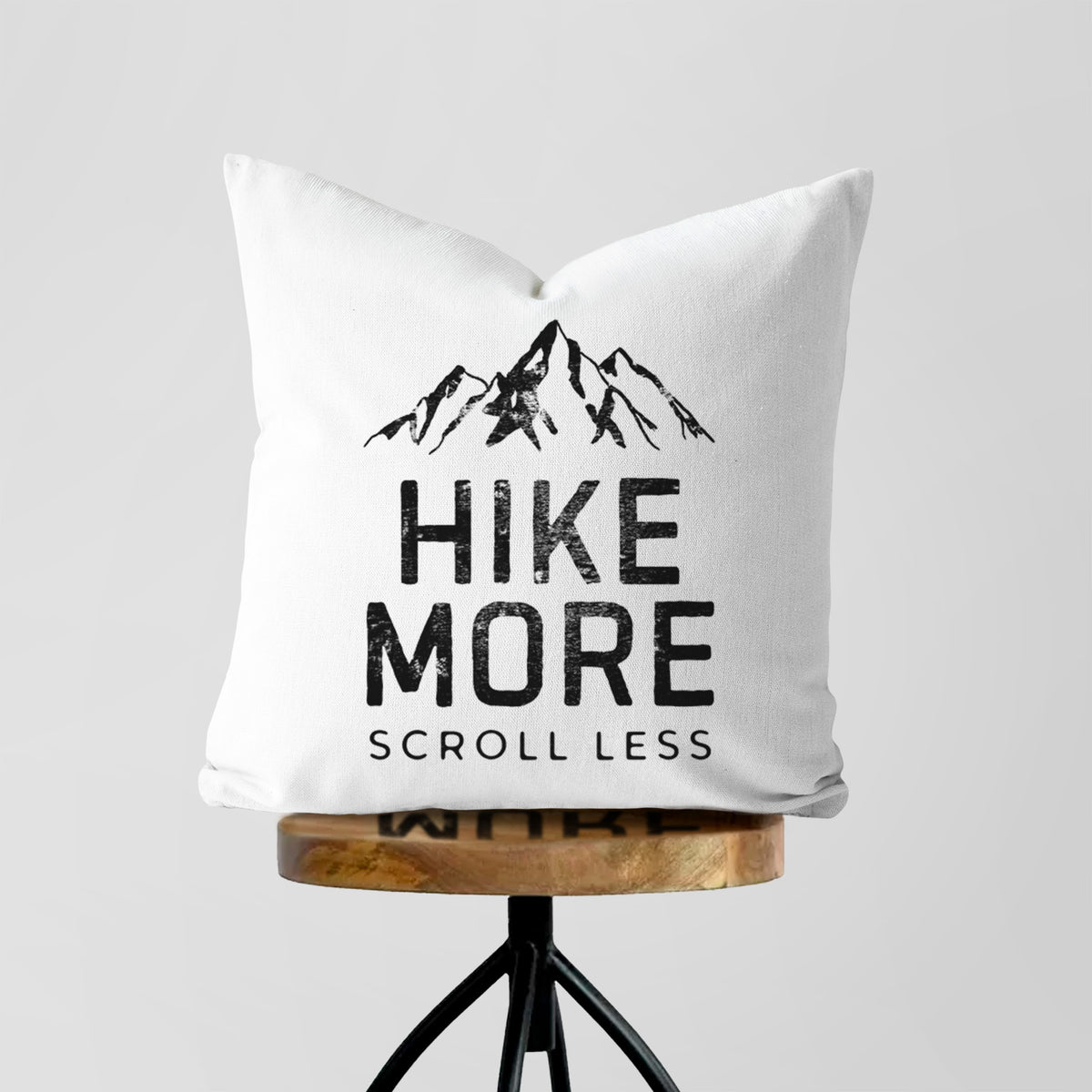 White pillow cover printed with black mountain peaks above the bold words Hike More, Scroll Less, on a wooden stool.