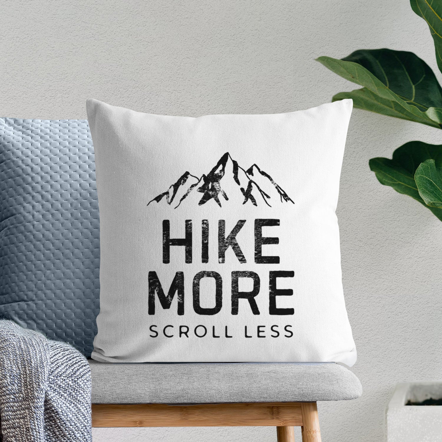 White pillow cover printed with black mountain peaks above the bold words Hike More, Scroll Less, on a bench beside plants.