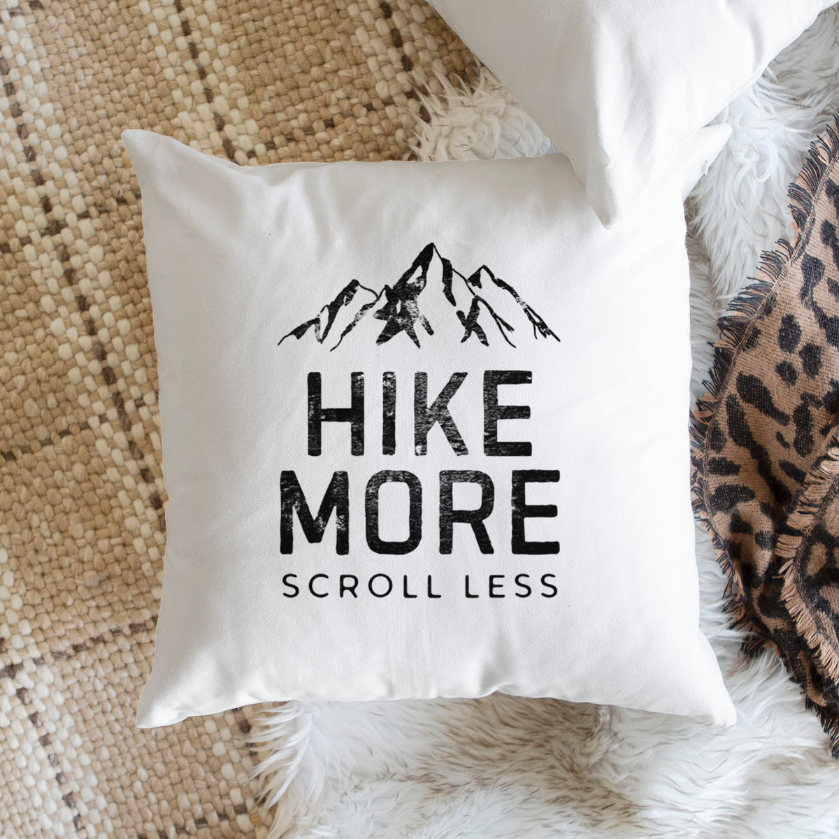White pillow cover printed with black mountain peaks above the bold words Hike More, Scroll Less, on a woven chair.
