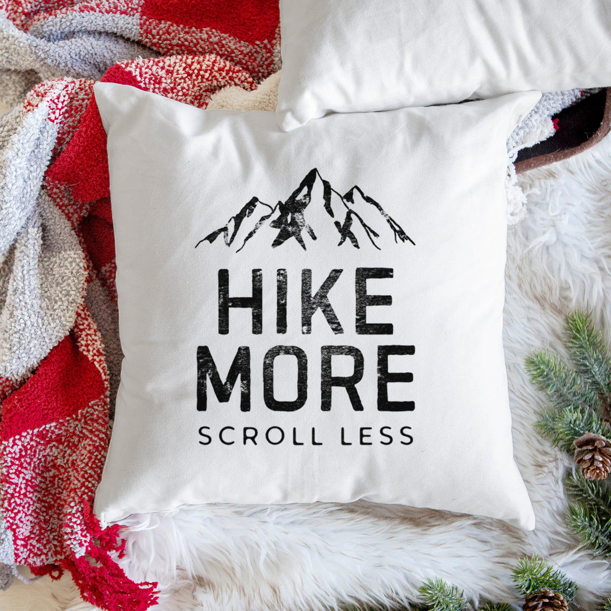 White pillow cover printed with black mountain peaks above the bold words Hike More, Scroll Less, on a woven chair.