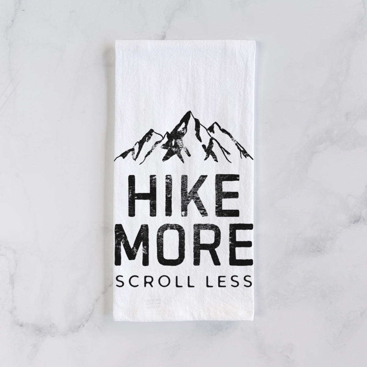 White cotton tea towel laid flat on marble, printed with black mountain peaks above the bold words Hike More, Scroll Less.