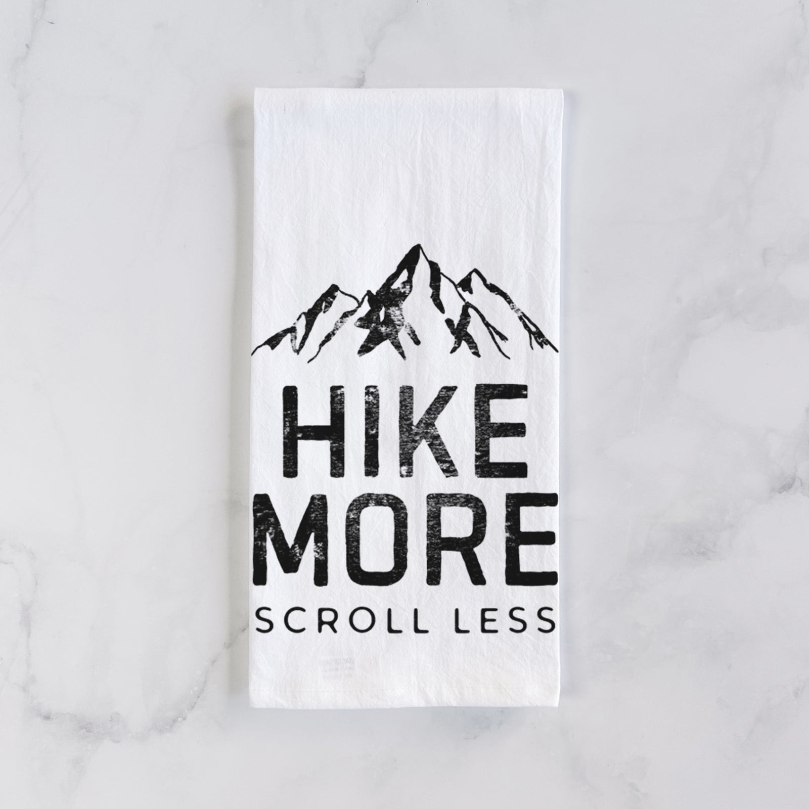 White cotton tea towel printed with black mountain peaks above the bold words Hike More, Scroll Less, on a marble counter beside a round wooden cutting board and greenery.