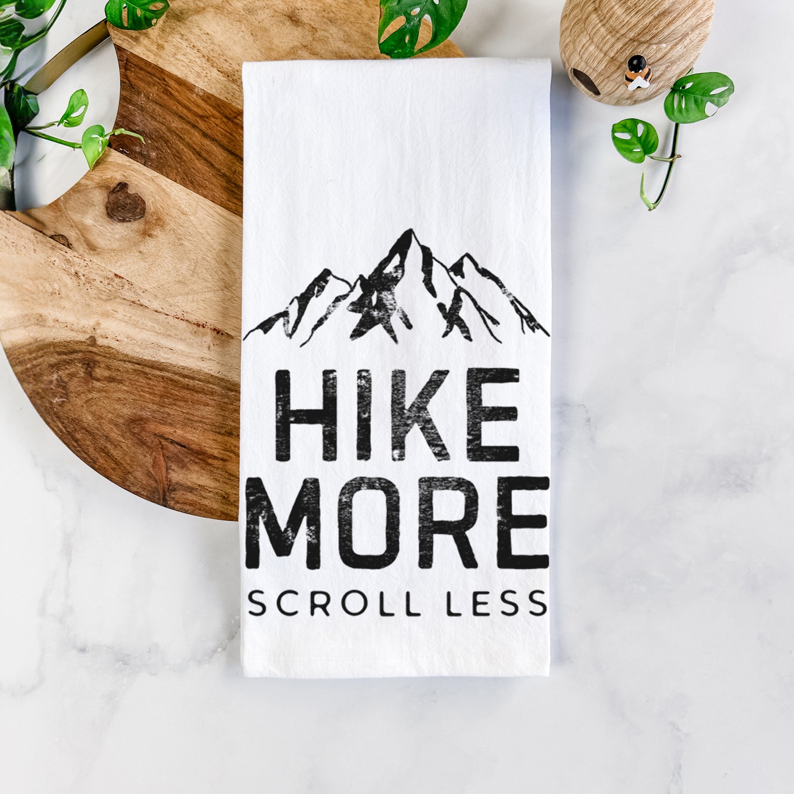White cotton tea towel printed with black mountain peaks above the bold words Hike More, Scroll Less, on a marble counter beside a round wooden cutting board and greenery.