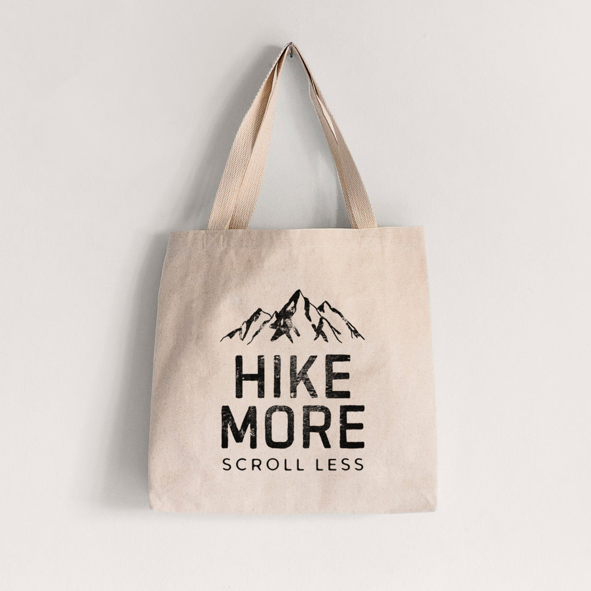 Natural canvas tote bag printed with black mountain peaks above the bold words Hike More, Scroll Less.