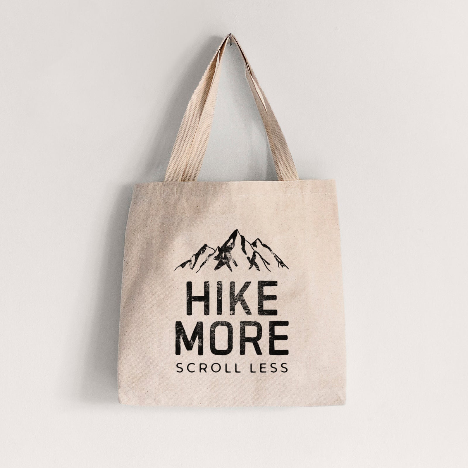 Natural canvas tote bag printed with black mountain peaks above the bold words Hike More, Scroll Less.
