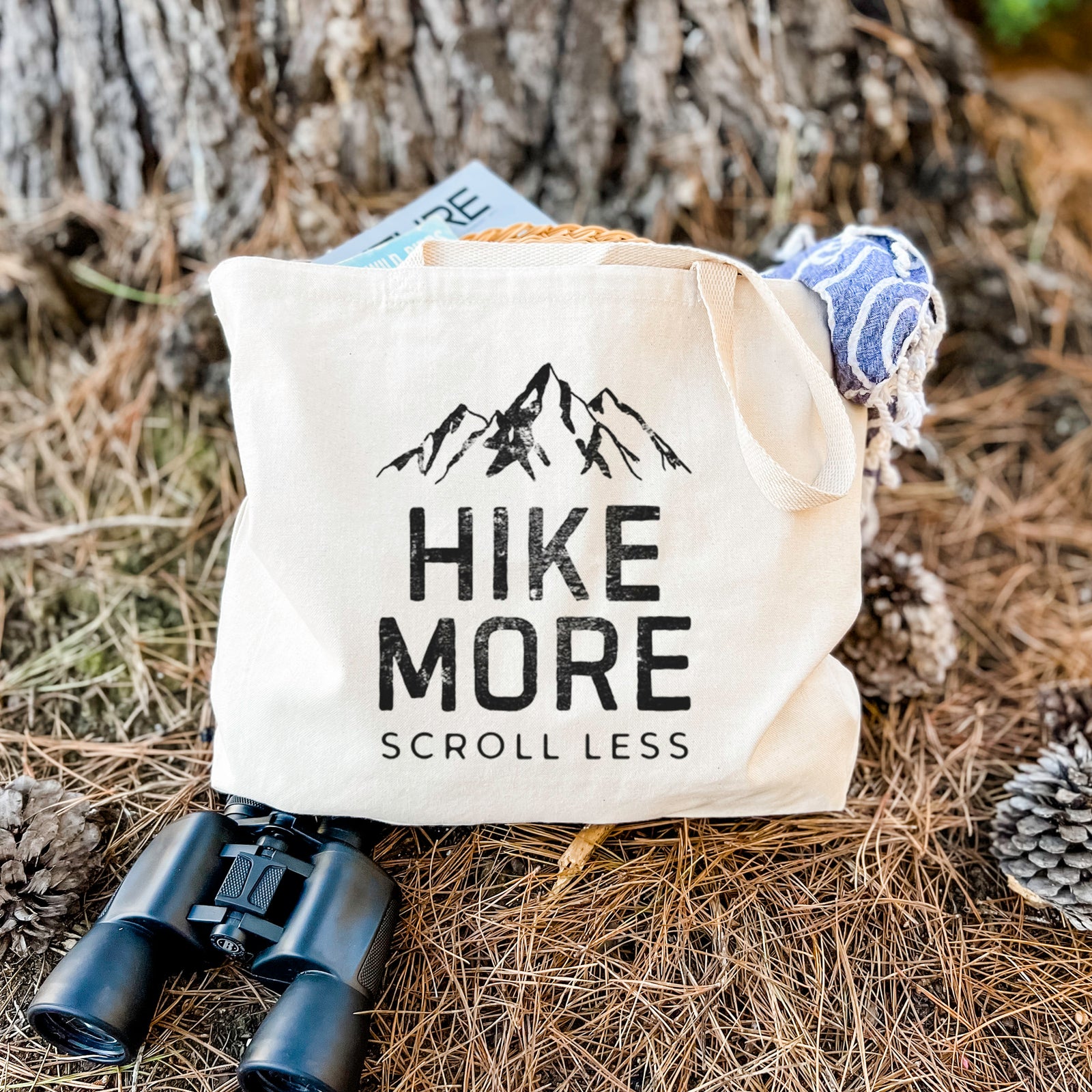 Natural canvas tote bag printed with black mountain peaks above the bold words Hike More, Scroll Less.
