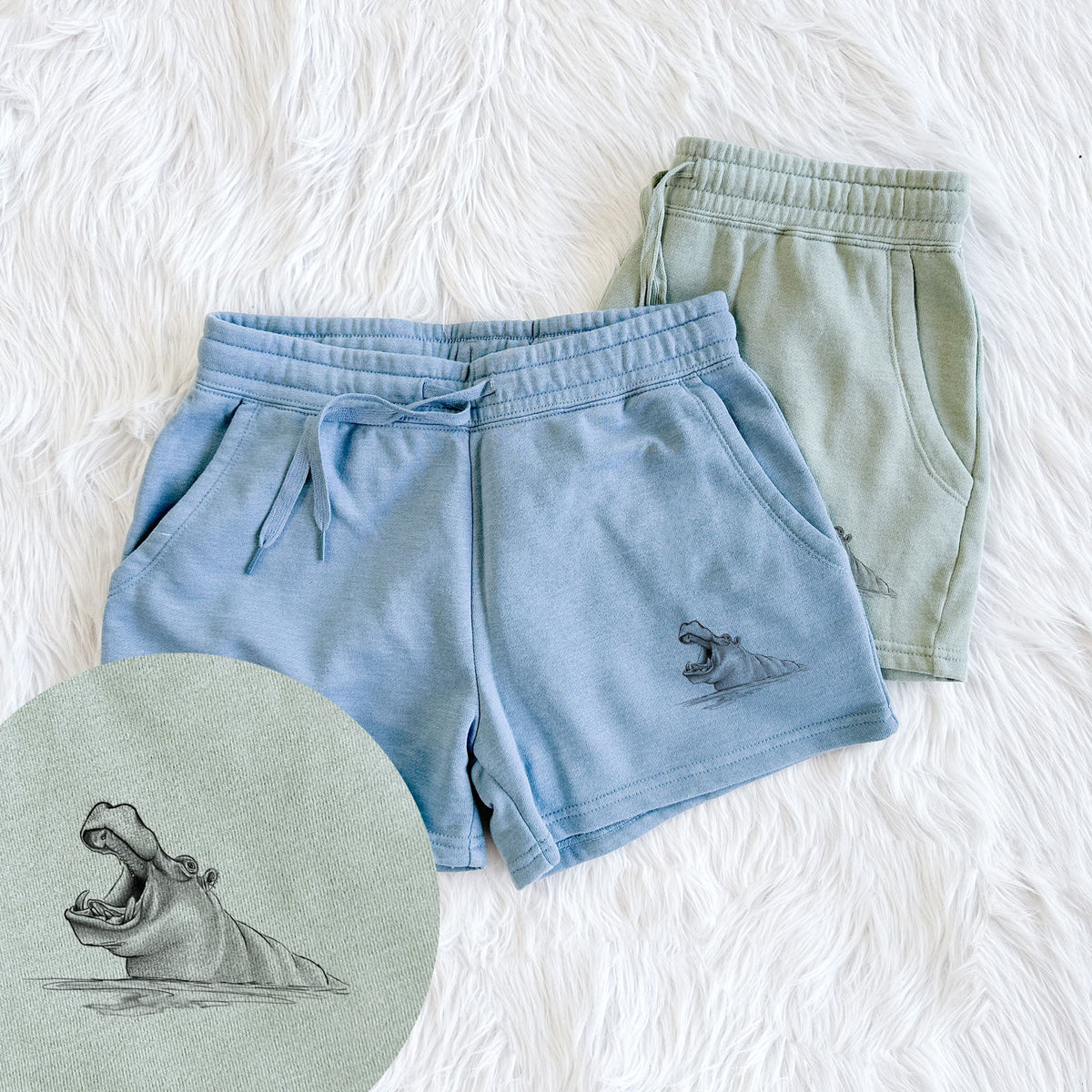 Two pairs of Cali Wave shorts in different colors printed with a black ink illustration of a hippo yawning with its mouth wide open, half submerged in water, with a close-up of the print.