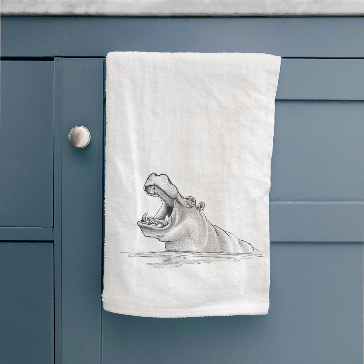 White hand towel printed with a black ink illustration of a hippo yawning with its mouth wide open, half submerged in water, hanging on a cabinet door.