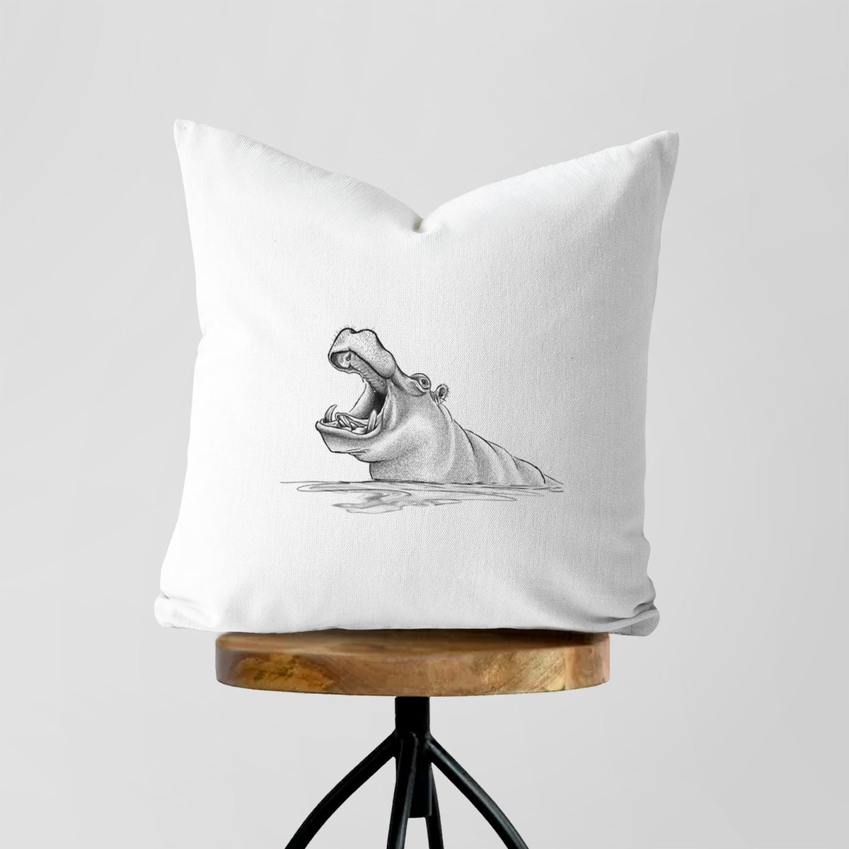 White pillow cover printed with a black ink illustration of a hippo yawning with its mouth wide open, half submerged in water, on a wooden stool.