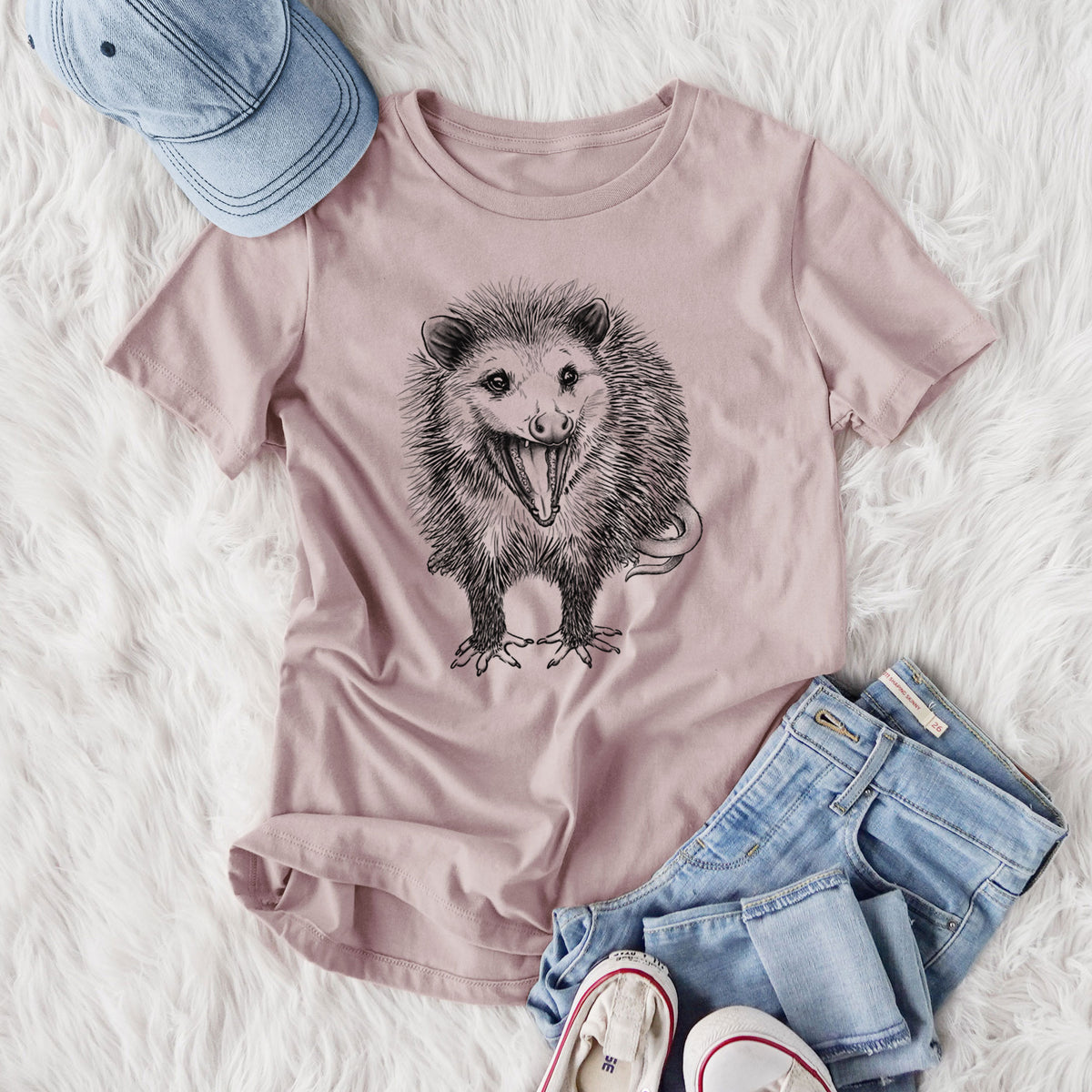 Women's relaxed fit cotton t-shirt printed with a detailed black ink illustration of an opossum standing and facing forward, mouth open wide in a hiss, with shaggy fur and its tail curled behind, styled flat on a white faux fur rug with jeans.