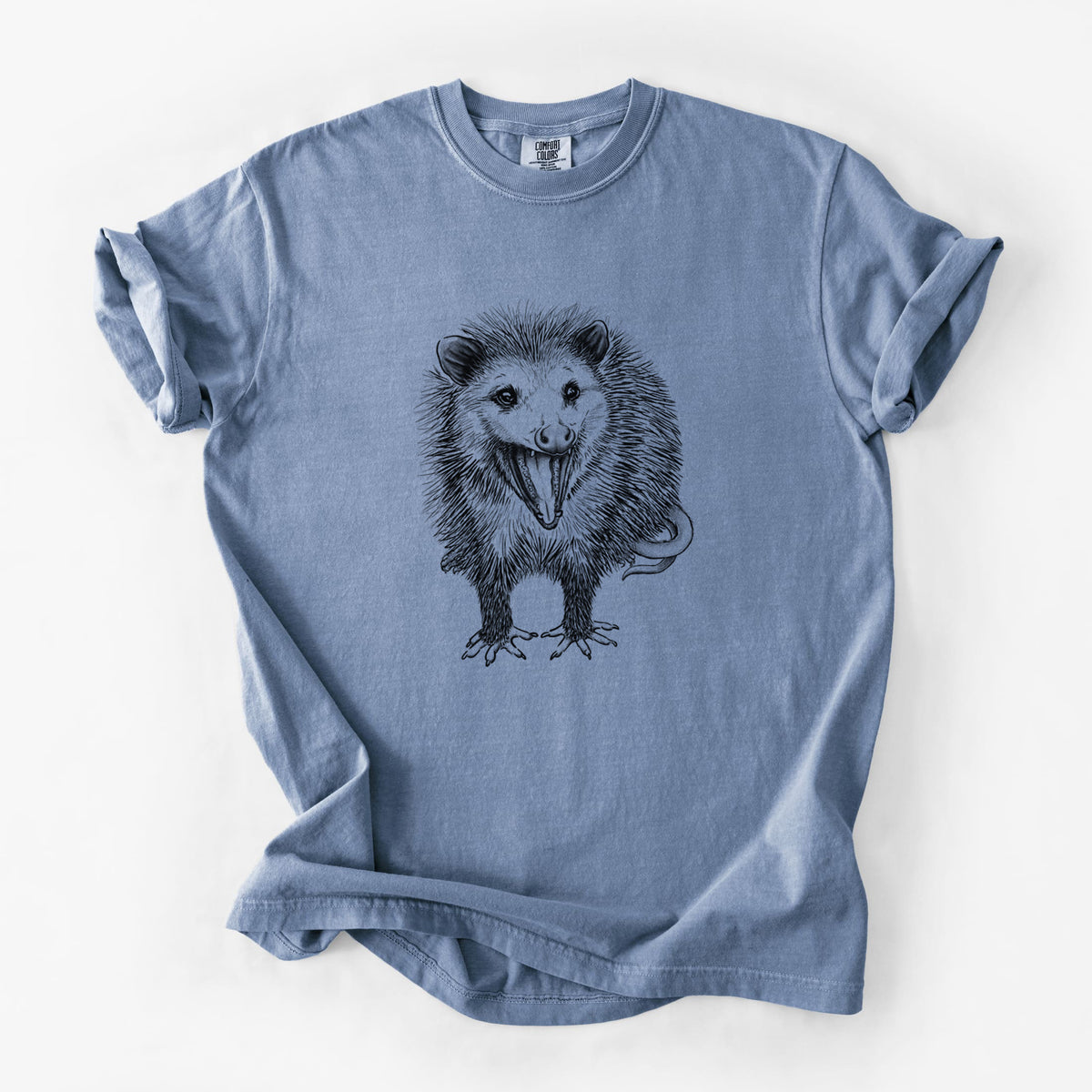 Blue jean Comfort Colors t-shirt laid flat, printed with a detailed black ink illustration of an opossum standing and facing forward, mouth open wide in a hiss, with shaggy fur and its tail curled behind.