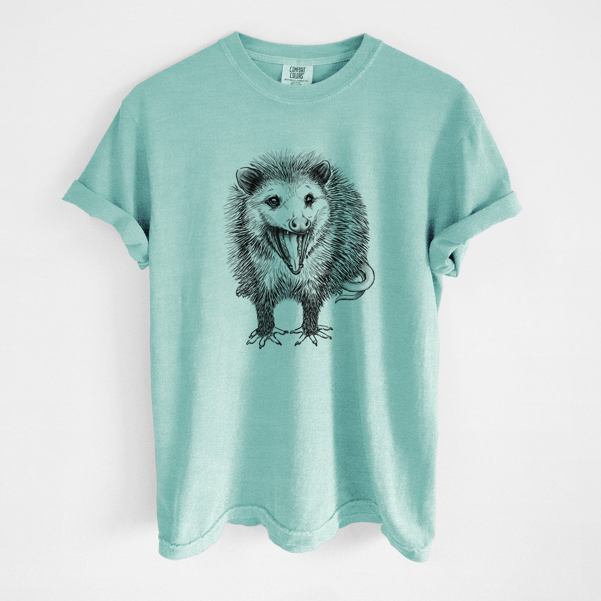 Chalky mint Comfort Colors t-shirt laid flat, printed with a detailed black ink illustration of an opossum standing and facing forward, mouth open wide in a hiss, with shaggy fur and its tail curled behind.
