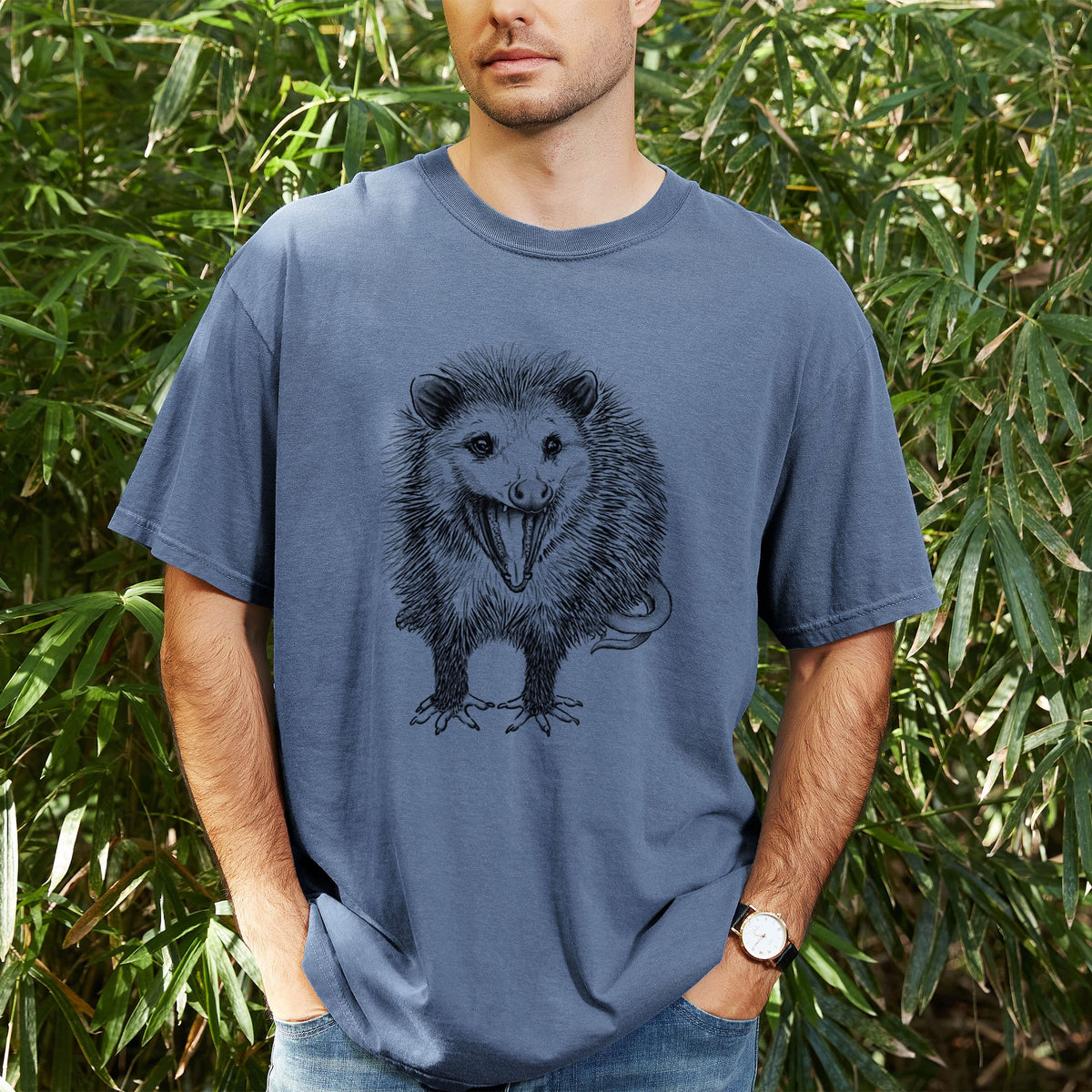 Man wearing a Comfort Colors t-shirt printed with a detailed black ink illustration of an opossum standing and facing forward, mouth open wide in a hiss, with shaggy fur and its tail curled behind, outdoors.