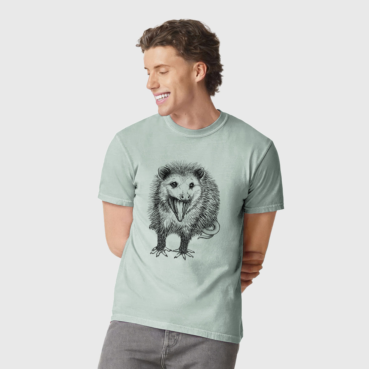 Man wearing a Comfort Colors t-shirt printed with a detailed black ink illustration of an opossum standing and facing forward, mouth open wide in a hiss, with shaggy fur and its tail curled behind.