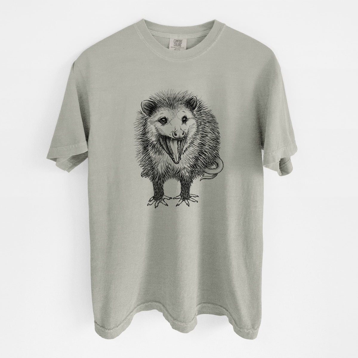 Sandstone beige Comfort Colors t-shirt laid flat, printed with a detailed black ink illustration of an opossum standing and facing forward, mouth open wide in a hiss, with shaggy fur and its tail curled behind.