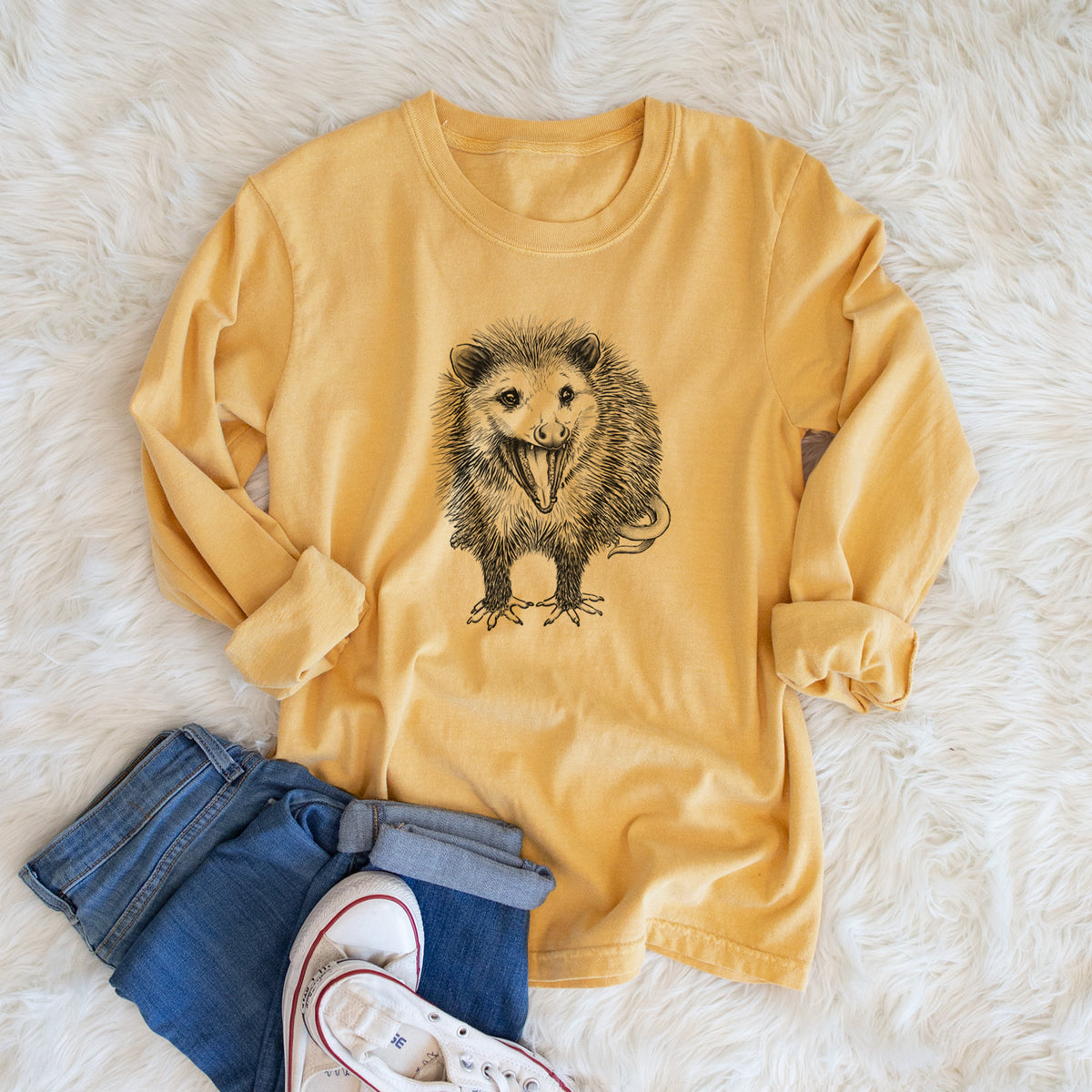 Golden wheat yellow Comfort Colors long sleeve shirt printed with a detailed black ink illustration of an opossum standing and facing forward, mouth open wide in a hiss, with shaggy fur and its tail curled behind, shown in a lifestyle photo.