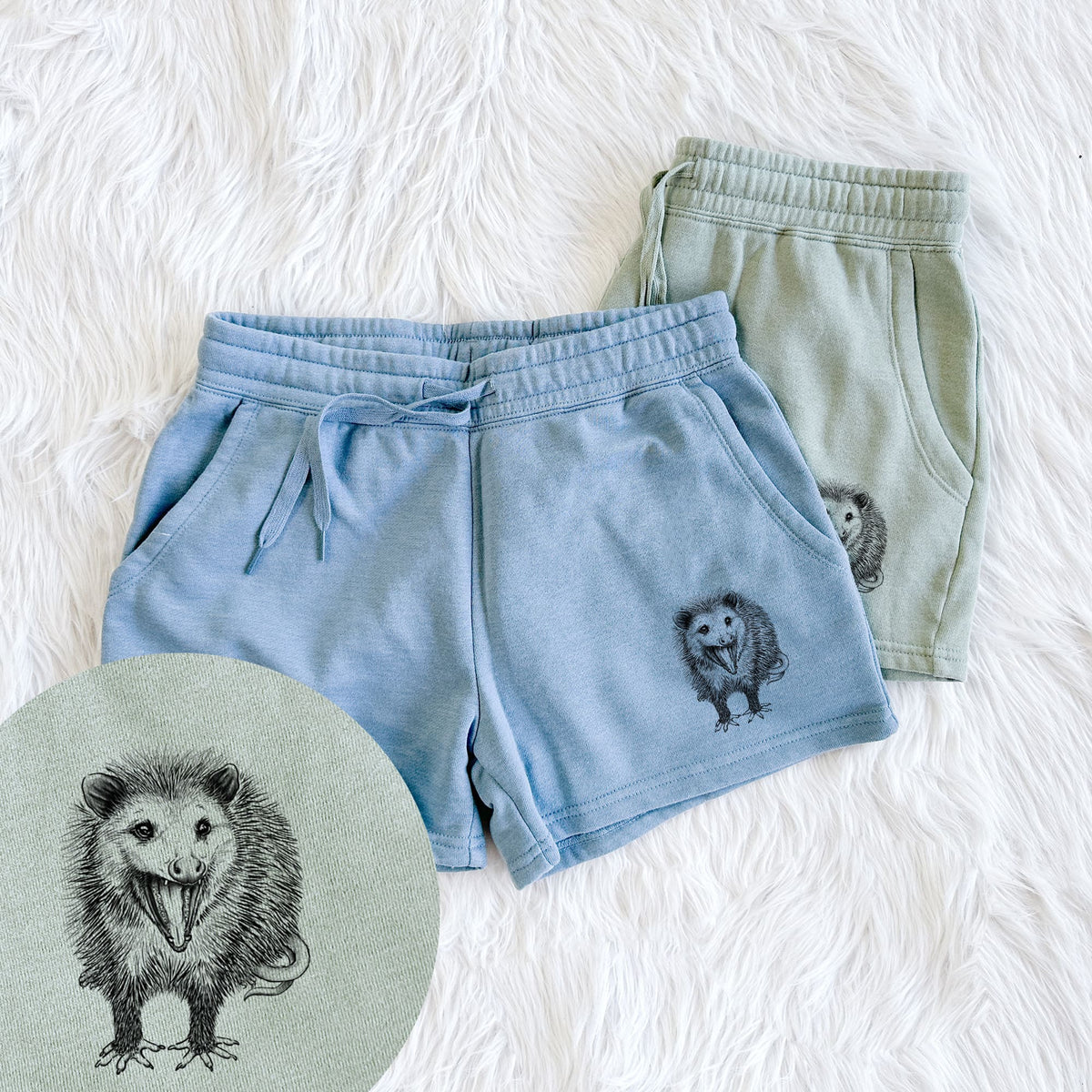 Two pairs of Cali Wave shorts in different colors printed with a detailed black ink illustration of an opossum standing and facing forward, mouth open wide in a hiss, with shaggy fur and its tail curled behind, with a close-up of the print.