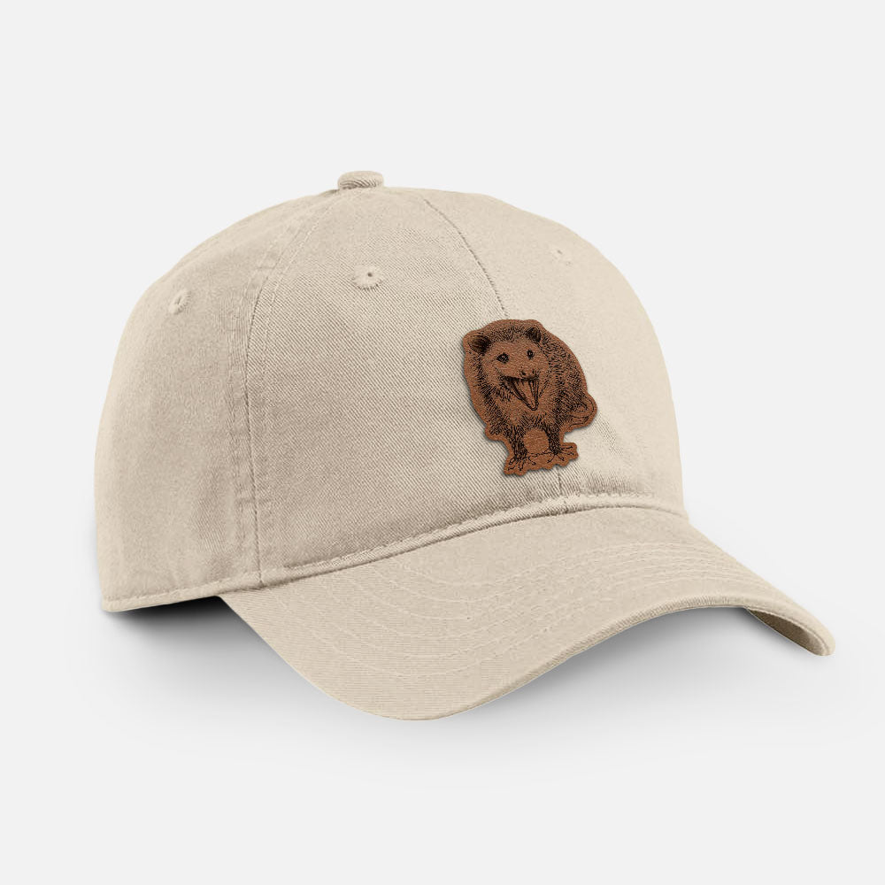 Stone cotton baseball hat with a detailed black ink illustration of an opossum standing and facing forward, mouth open wide in a hiss, with shaggy fur and its tail curled behind on the front.