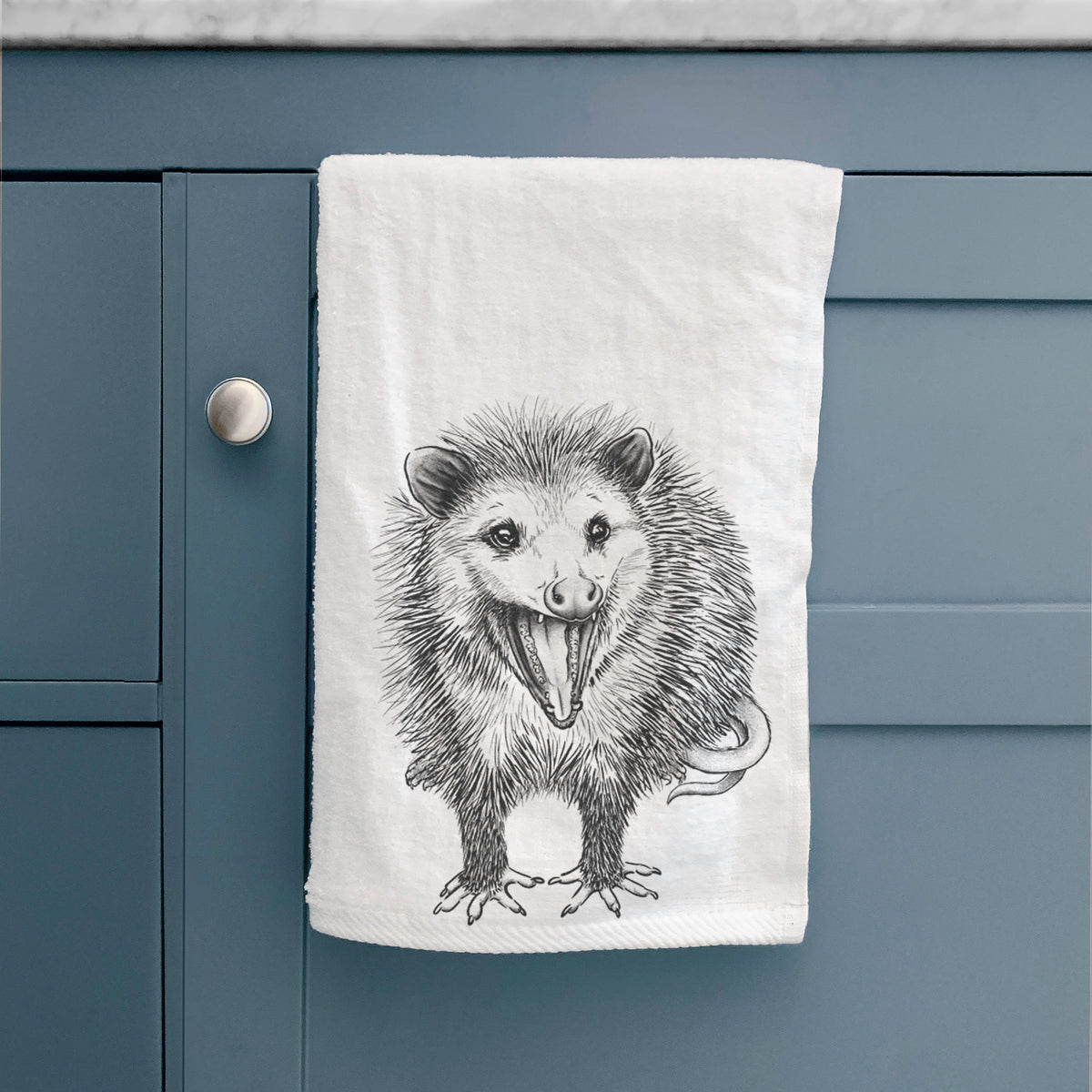 White hand towel printed with a detailed black ink illustration of an opossum standing and facing forward, mouth open wide in a hiss, with shaggy fur and its tail curled behind, hanging on a cabinet door.