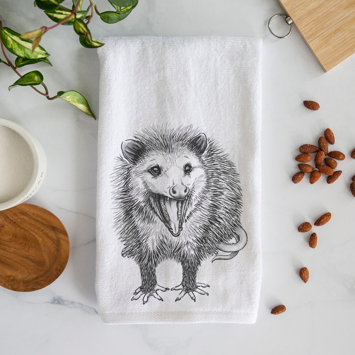 White hand towel printed with a detailed black ink illustration of an opossum standing and facing forward, mouth open wide in a hiss, with shaggy fur and its tail curled behind.