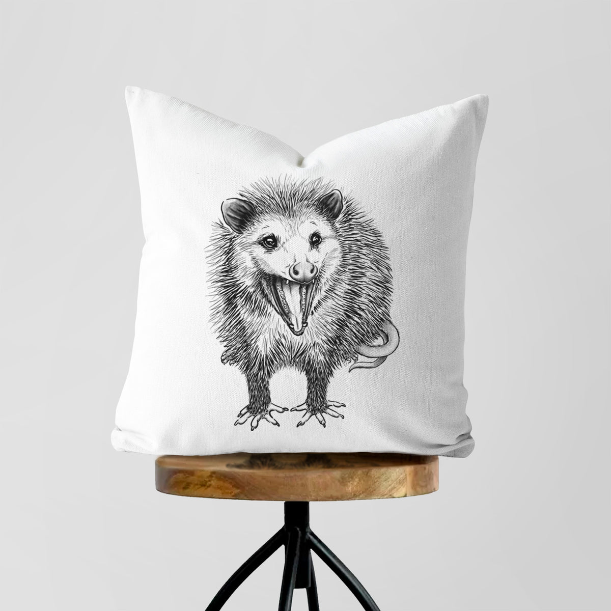 White pillow cover printed with a detailed black ink illustration of an opossum standing and facing forward, mouth open wide in a hiss, with shaggy fur and its tail curled behind, on a wooden stool.