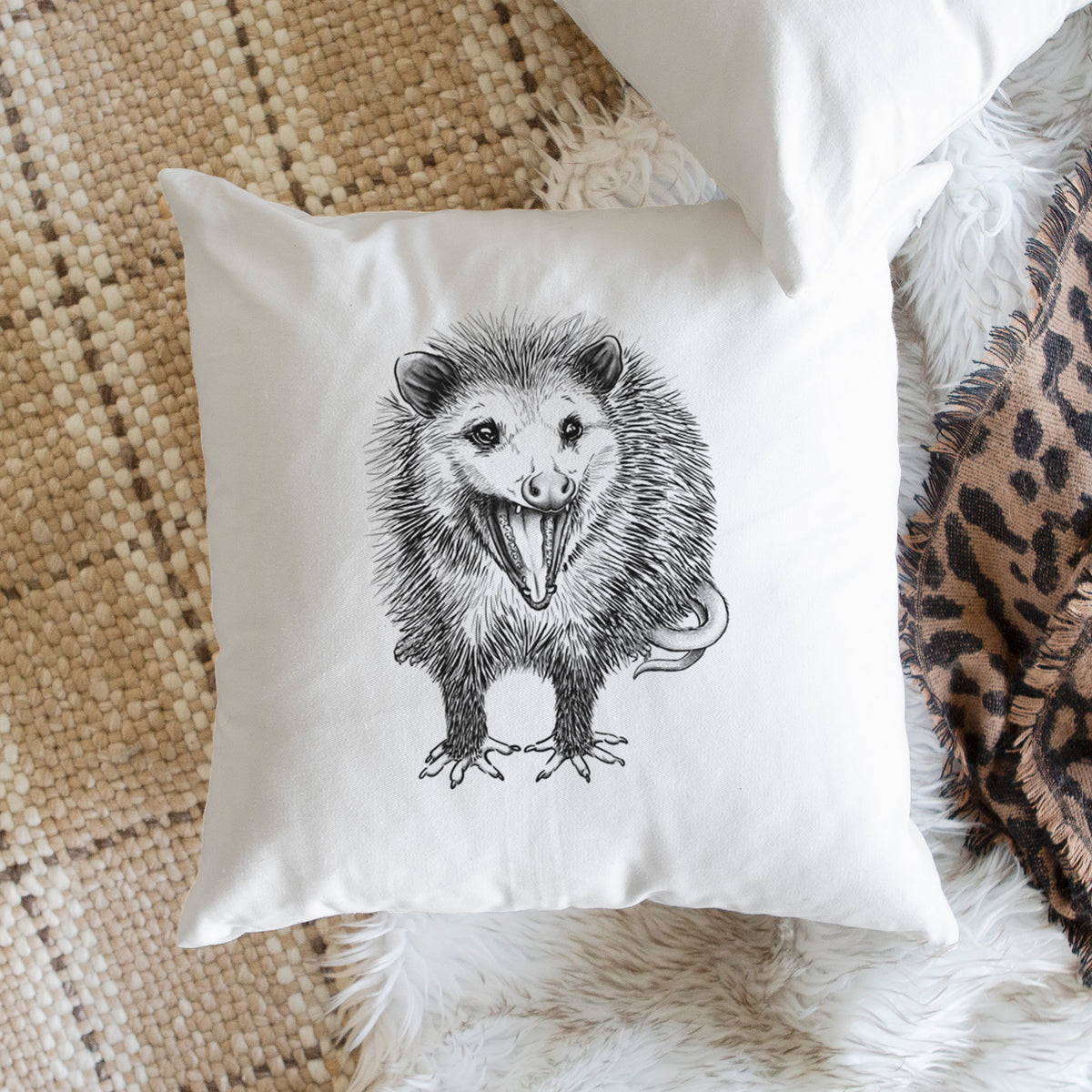 White pillow cover printed with a detailed black ink illustration of an opossum standing and facing forward, mouth open wide in a hiss, with shaggy fur and its tail curled behind, on a woven chair.