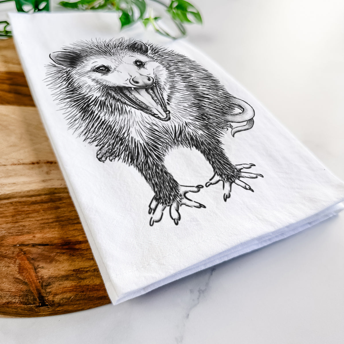 Close-up of a folded white tea towel on a wooden board, showing a detailed black ink illustration of an opossum standing and facing forward, mouth open wide in a hiss, with shaggy fur and its tail curled behind.
