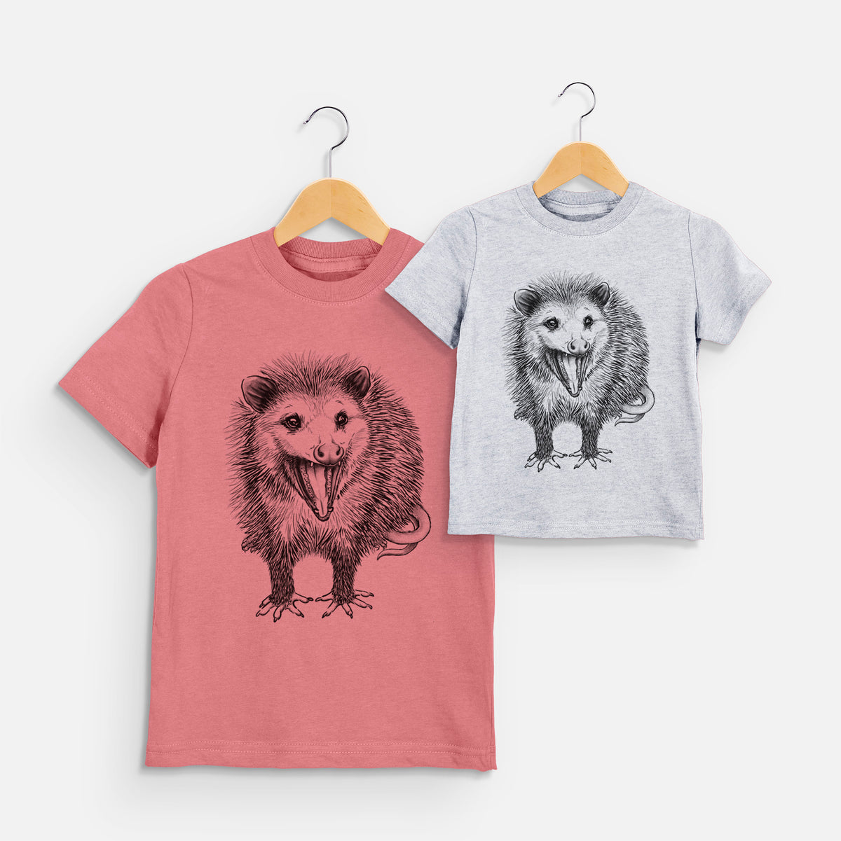 Two youth t-shirts in different colors on hangers, printed with a detailed black ink illustration of an opossum standing and facing forward, mouth open wide in a hiss, with shaggy fur and its tail curled behind.