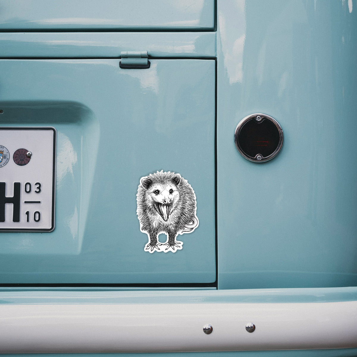 4 inch vinyl decal of a detailed black ink illustration of an opossum standing and facing forward, mouth open wide in a hiss, with shaggy fur and its tail curled behind on the back of a van.