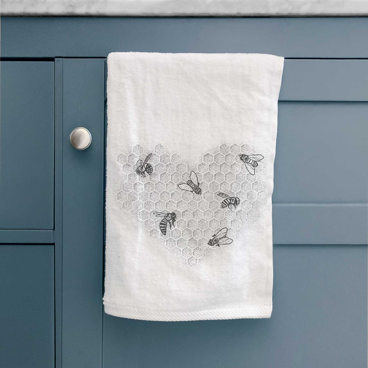 White hand towel printed with a black ink heart made of honeycomb cells with small bees crawling across it, hanging on a cabinet door.