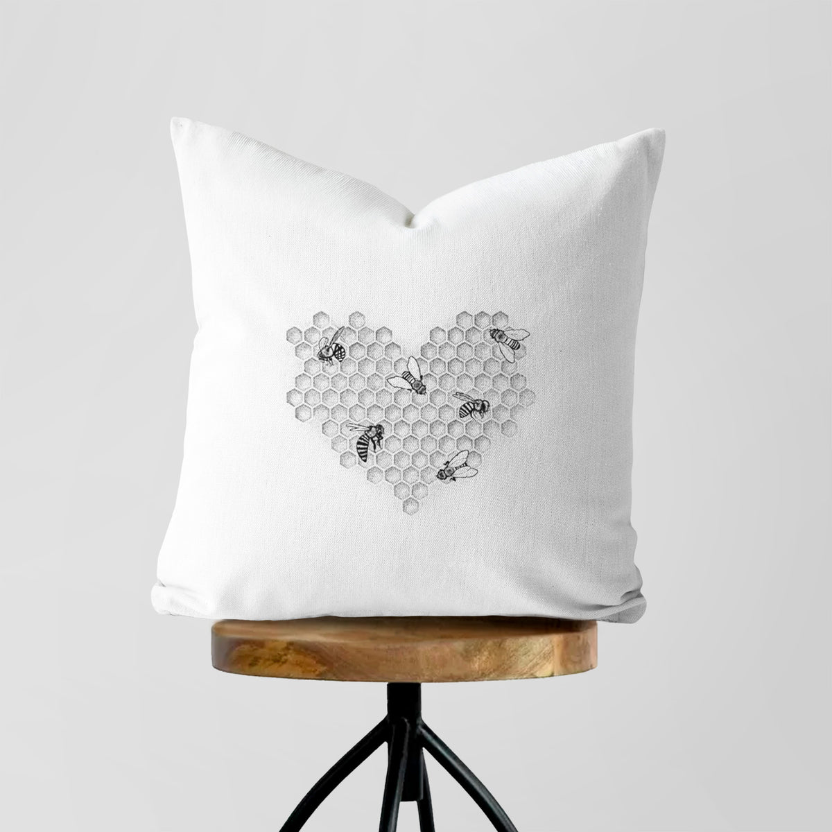 White pillow cover printed with a black ink heart made of honeycomb cells with small bees crawling across it, on a wooden stool.