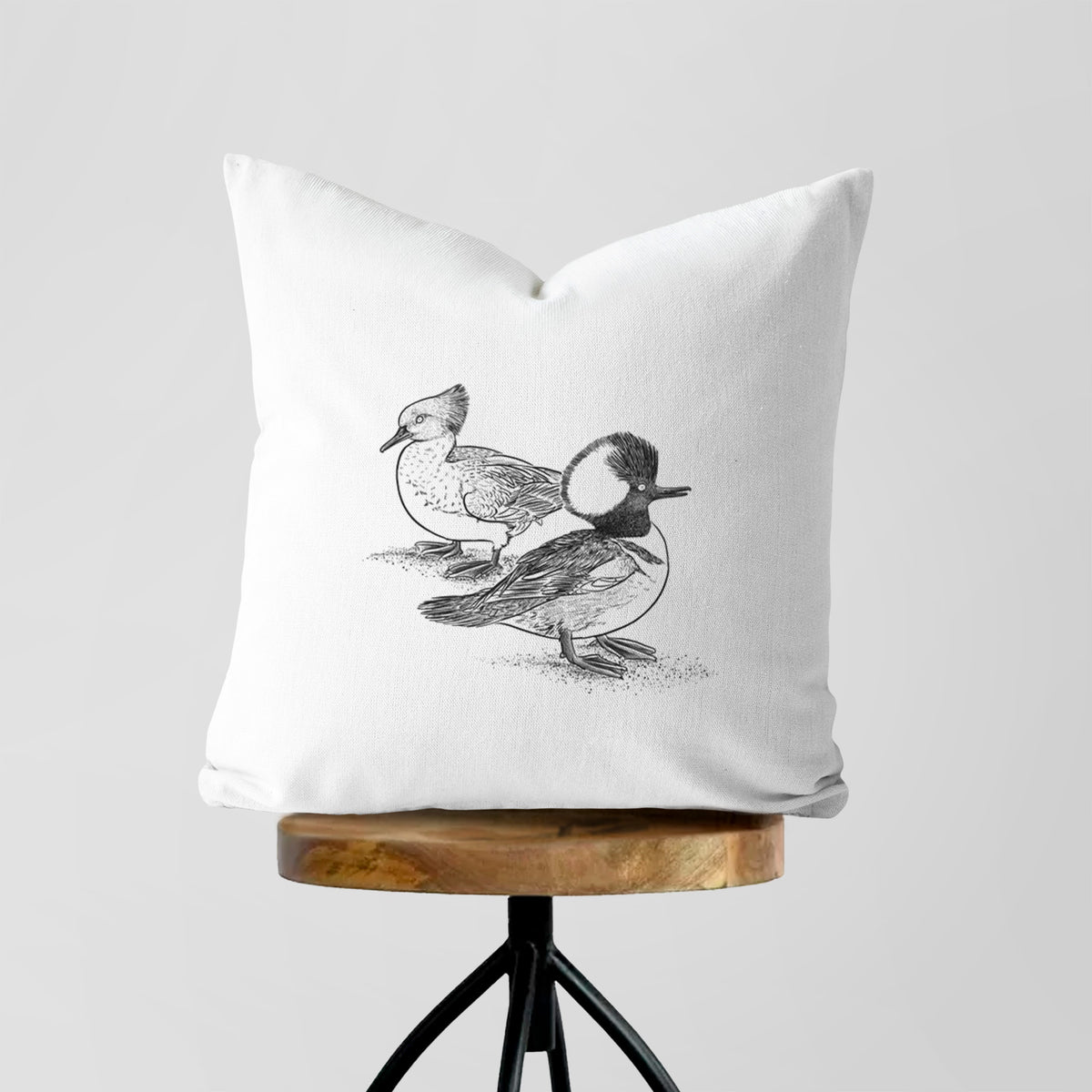 White pillow cover printed with a detailed black ink illustration of a pair of hooded mergansers, male and female, on a wooden stool.