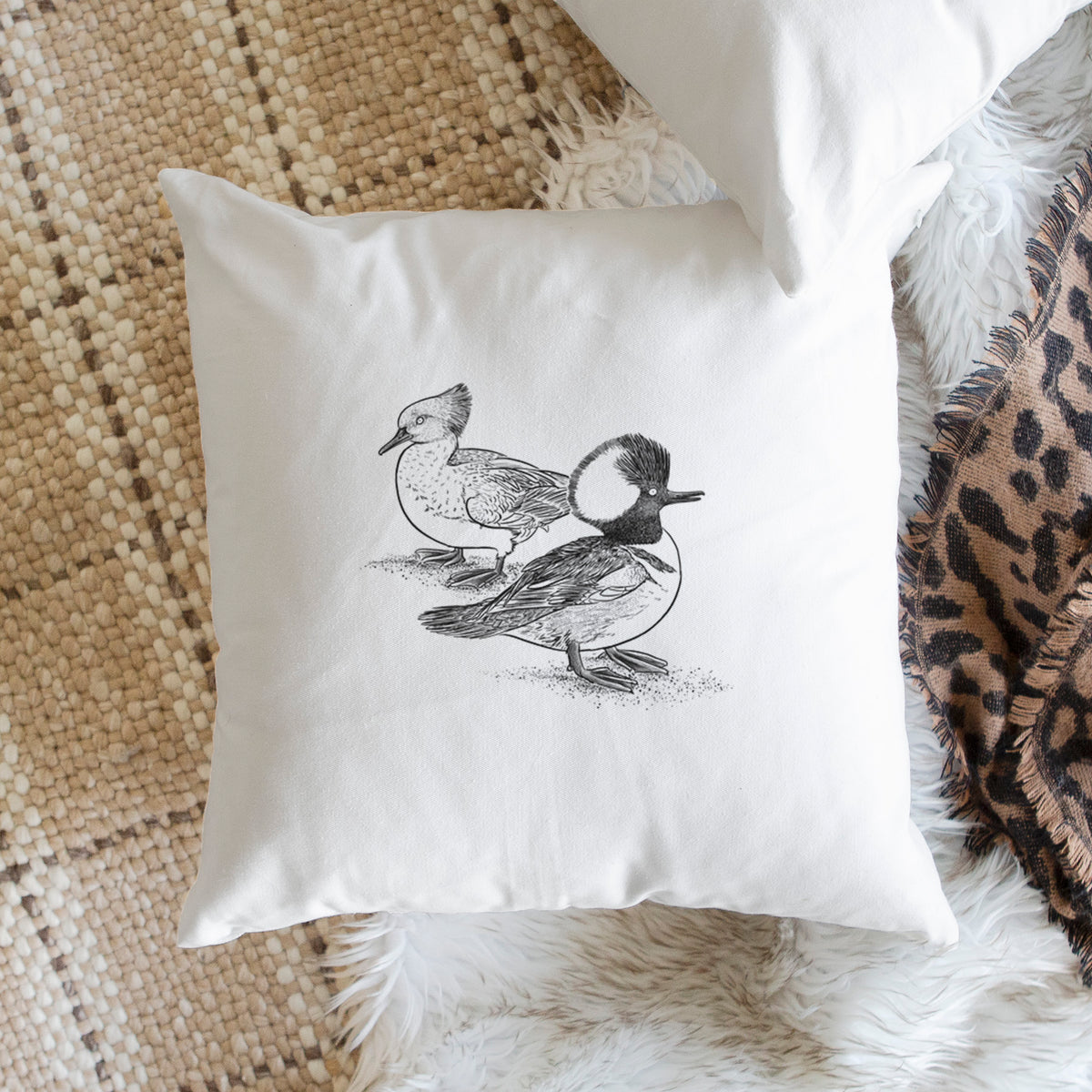 White pillow cover printed with a detailed black ink illustration of a pair of hooded mergansers, male and female, on a woven chair.