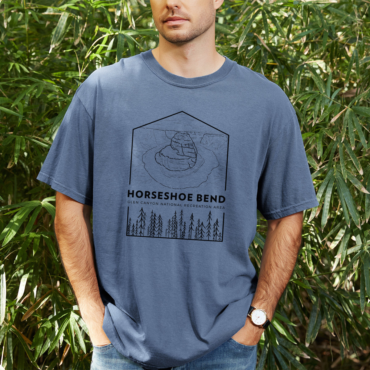 Man wearing a Comfort Colors t-shirt printed with a black line art badge of Horseshoe Bend above pine trees, with the words Horseshoe Bend, outdoors.