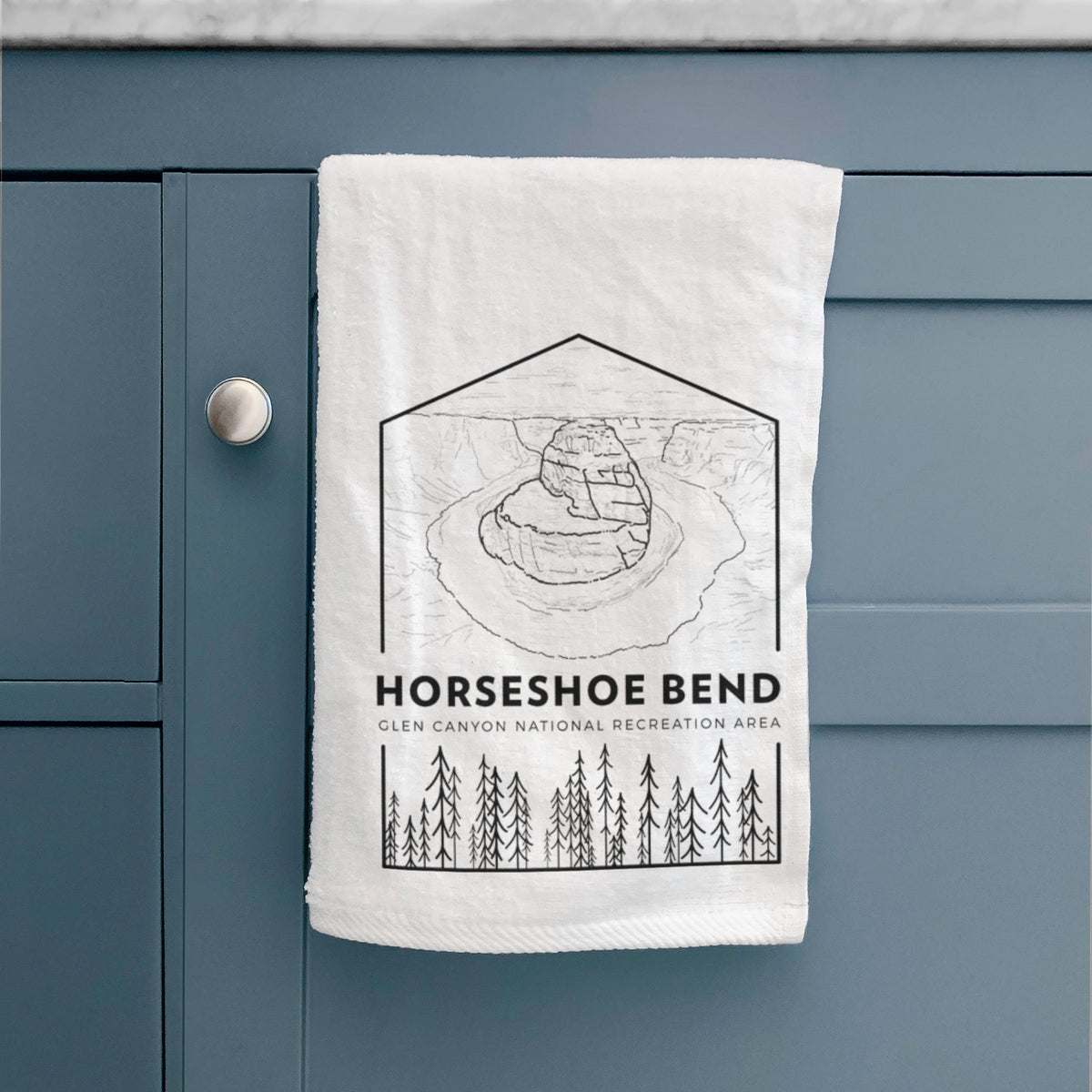 White hand towel printed with a black line art badge of Horseshoe Bend above pine trees, with the words Horseshoe Bend, hanging on a cabinet door.