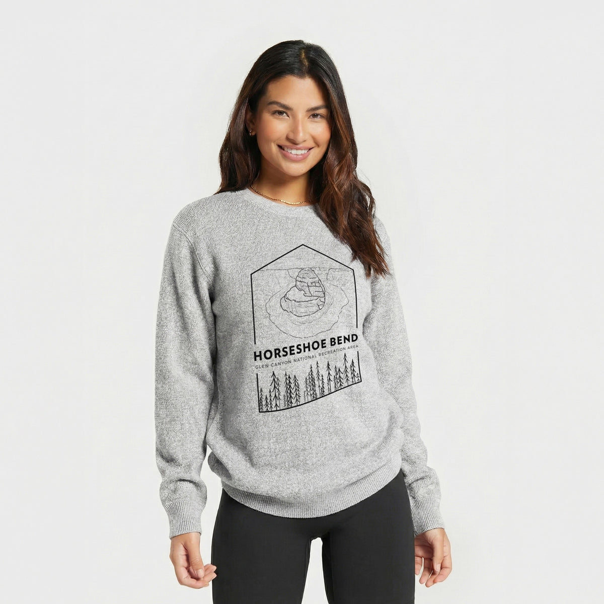 Woman wearing a knit sweatshirt printed with a black line art badge of Horseshoe Bend above pine trees, with the words Horseshoe Bend.