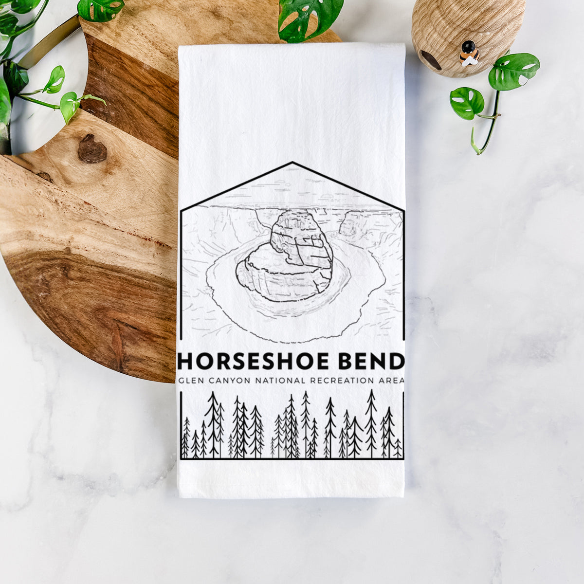 White cotton tea towel printed with a black line art badge of Horseshoe Bend above pine trees, with the words Horseshoe Bend, on a marble counter beside a round wooden cutting board and greenery.