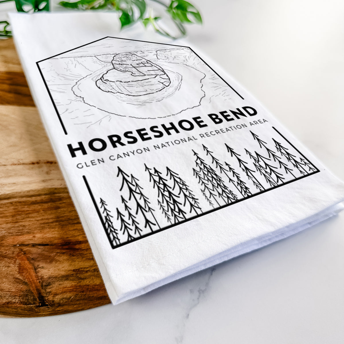 Close-up of a folded white tea towel on a wooden board, showing a black line art badge of Horseshoe Bend above pine trees, with the words Horseshoe Bend.