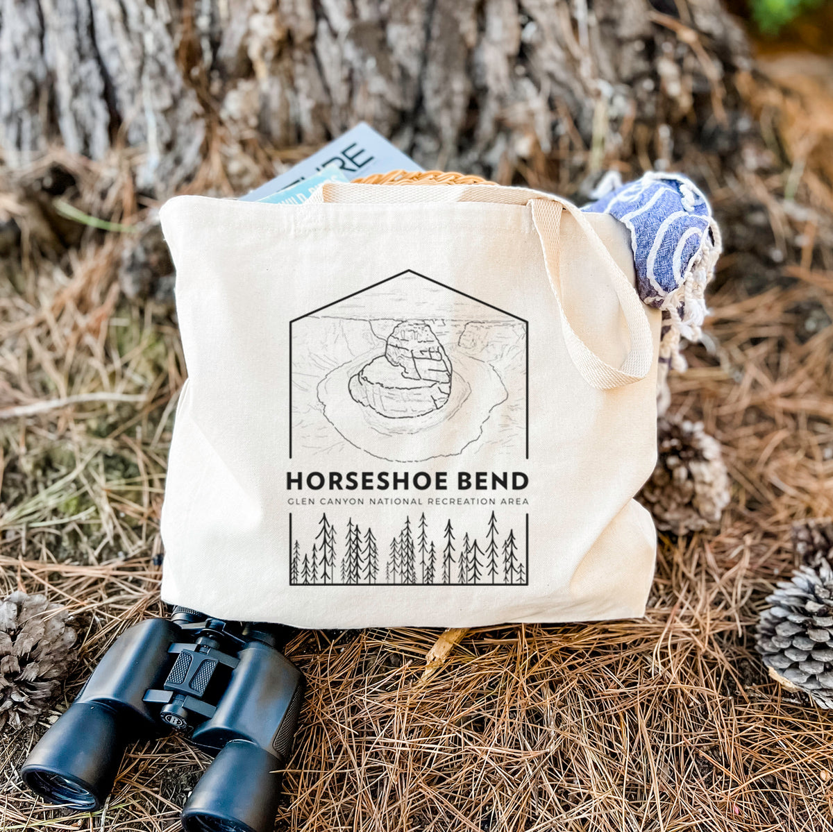 Canvas tote bag printed with a black line art badge of Horseshoe Bend above pine trees, with the words Horseshoe Bend, resting on dry grass beside a pair of binoculars.