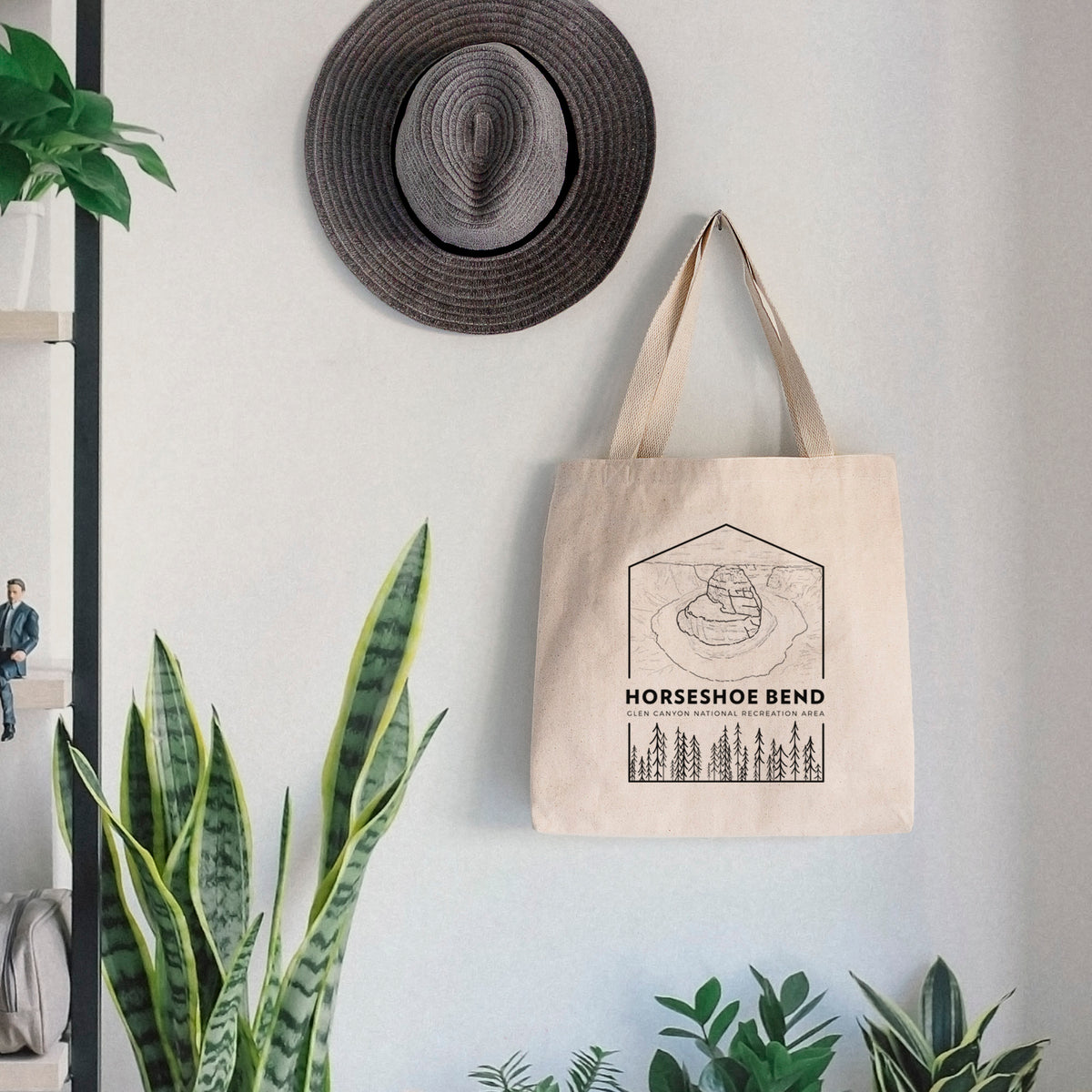 Canvas tote bag printed with a black line art badge of Horseshoe Bend above pine trees, with the words Horseshoe Bend, hanging on a wall hook below a hat, next to potted plants.