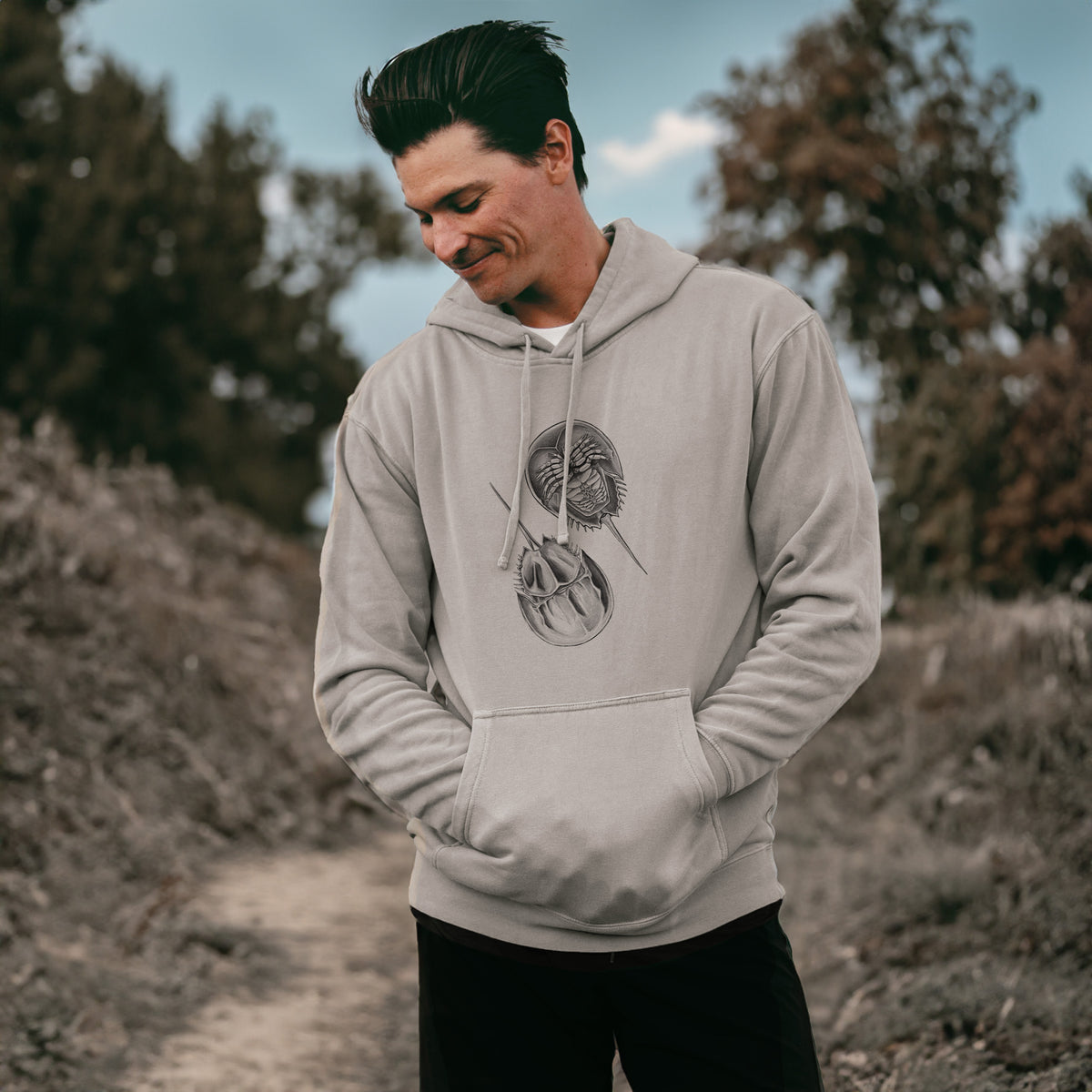 Man wearing a cement grey pigment dyed hoodie printed with a detailed black ink illustration of two horseshoe crabs.