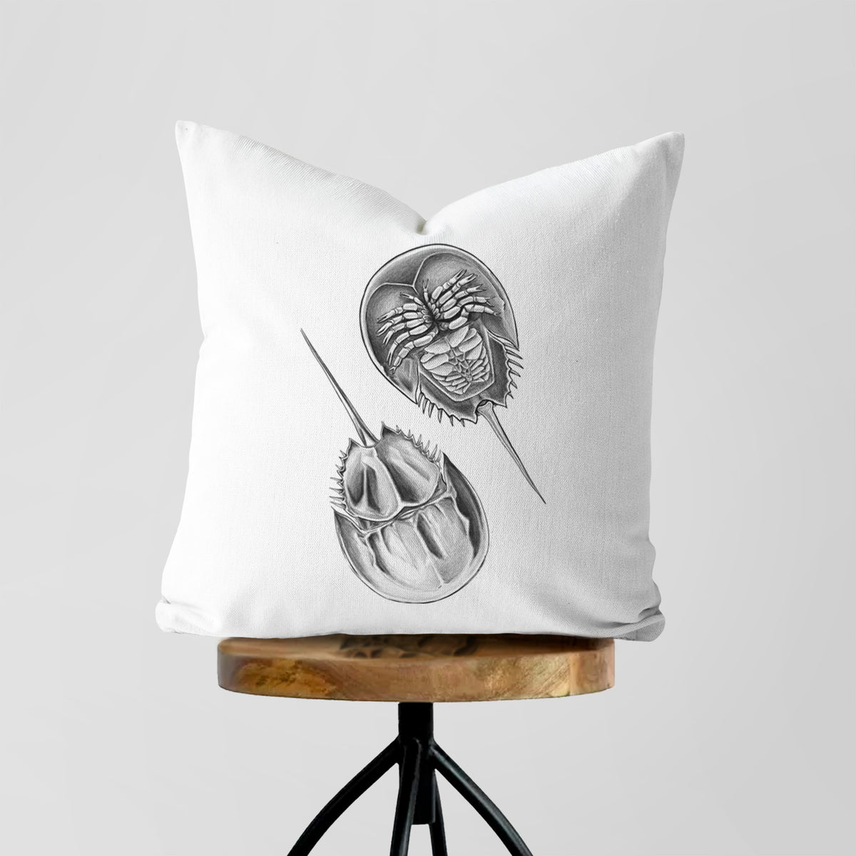 White pillow cover printed with a detailed black ink illustration of two horseshoe crabs, on a wooden stool.