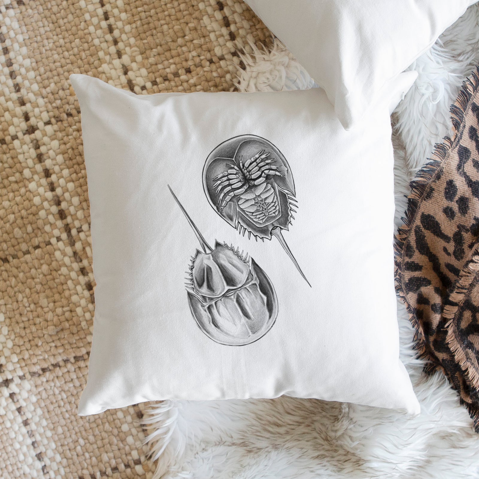 White pillow cover printed with a detailed black ink illustration of two horseshoe crabs, on a bench beside plants.
