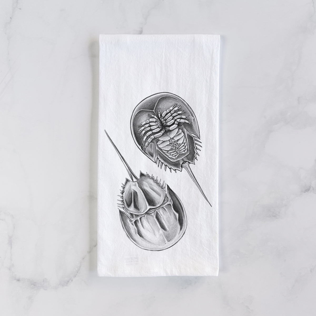 White cotton tea towel laid flat on marble, printed with a detailed black ink illustration of two horseshoe crabs.